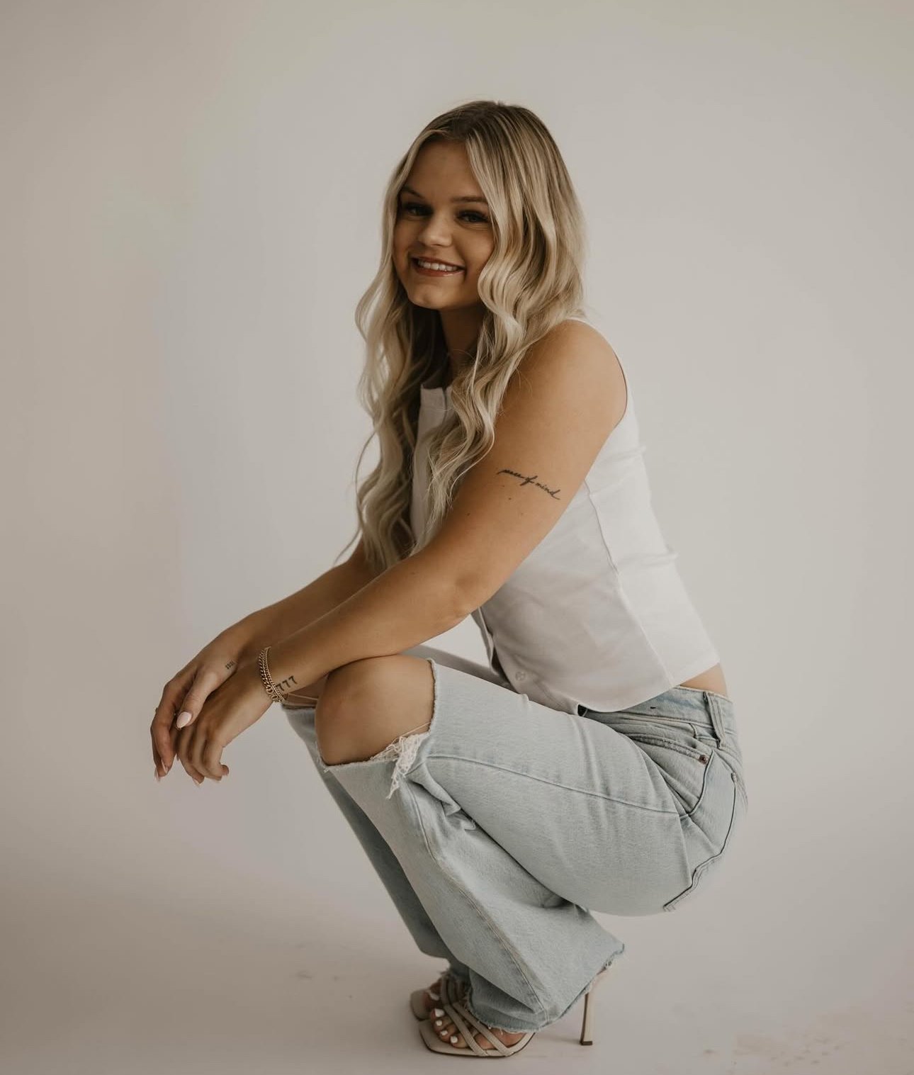 A young woman with long, wavy blonde hair, wearing a white sleeveless top and ripped light-wash jeans, smiling while squatting with one knee raised, against a plain neutral background.
