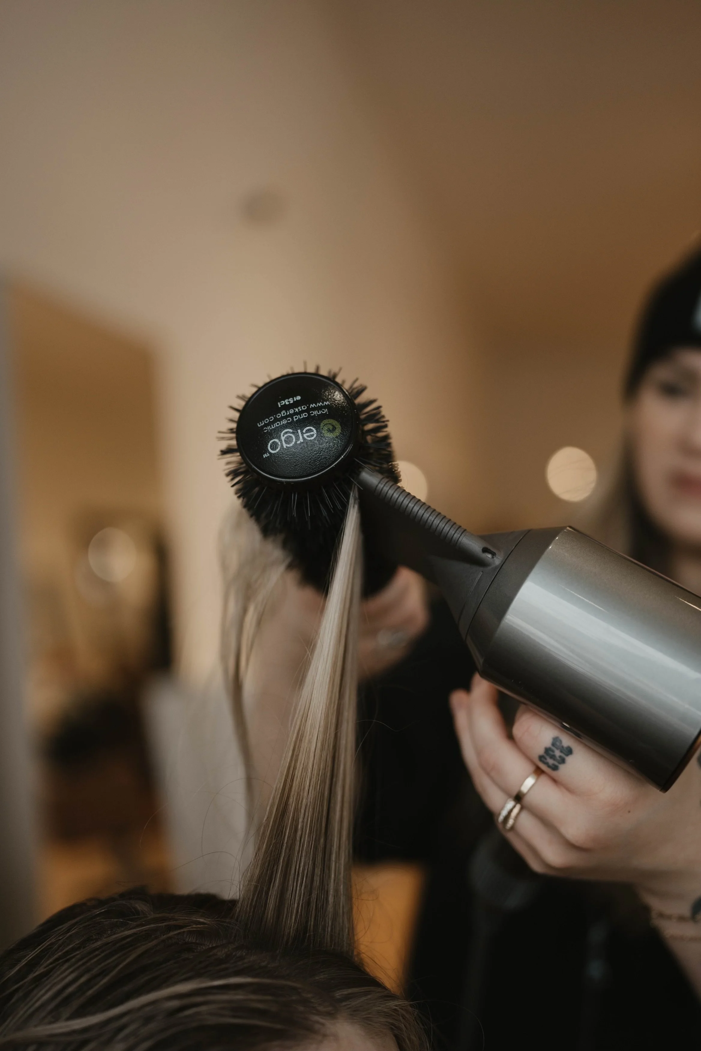 A person is blow-drying long, light brown hair with a black hairbrush attached to a hairdryer. The focus is on the hairdryer and brush, while the person's face is partially visible on the right side of the image.