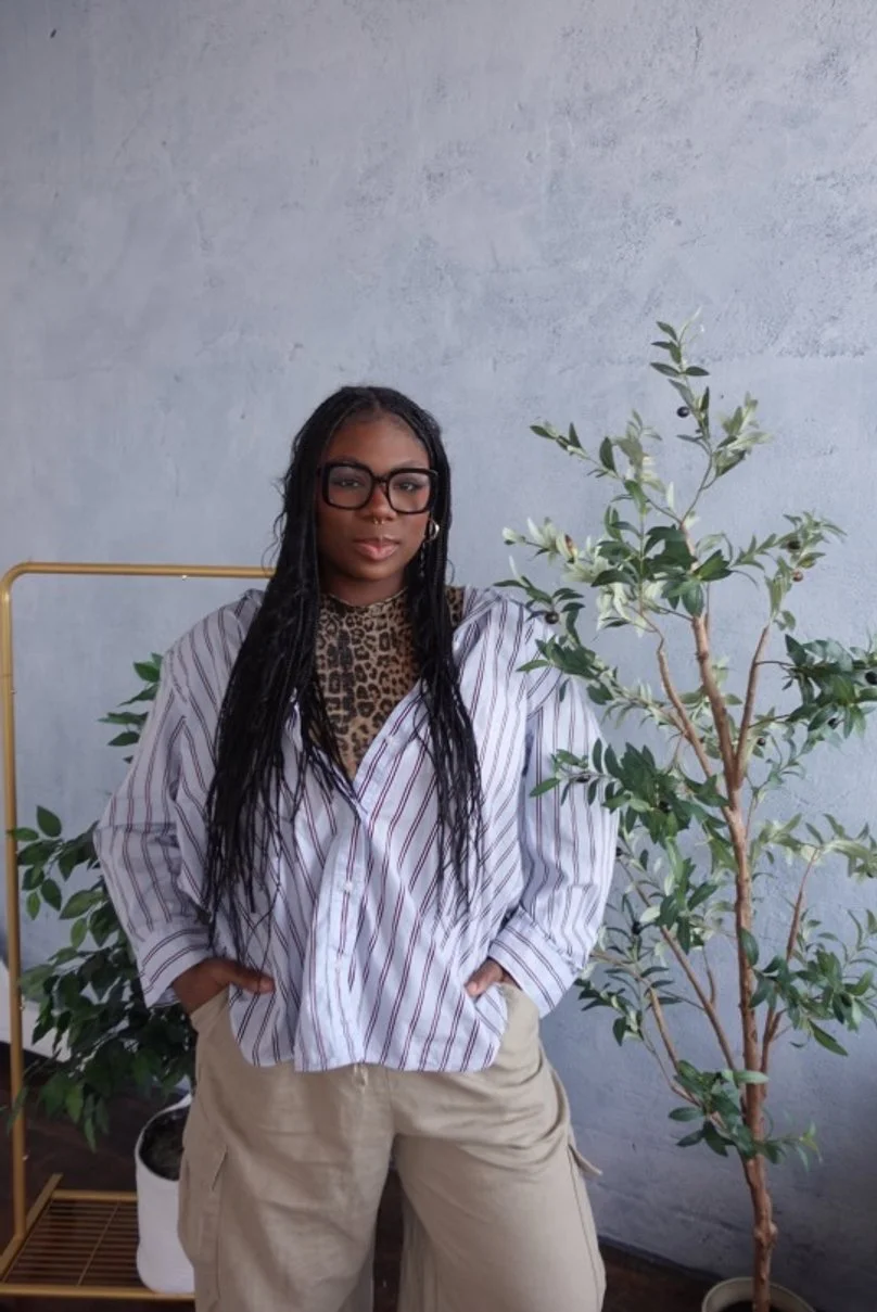 A woman with long braided hair, wearing glasses, a leopard print turtleneck, an oversized striped shirt, and beige cargo pants, standing with hands in pockets next to a plant against a light gray textured wall.