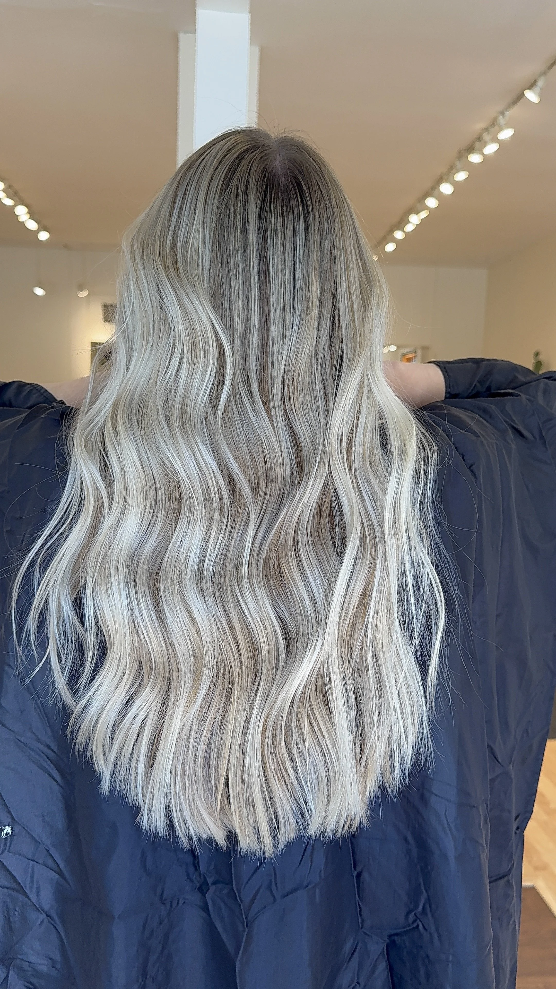 Person with long, wavy blonde hair, sitting in a salon or similar setting, wearing a dark jacket.