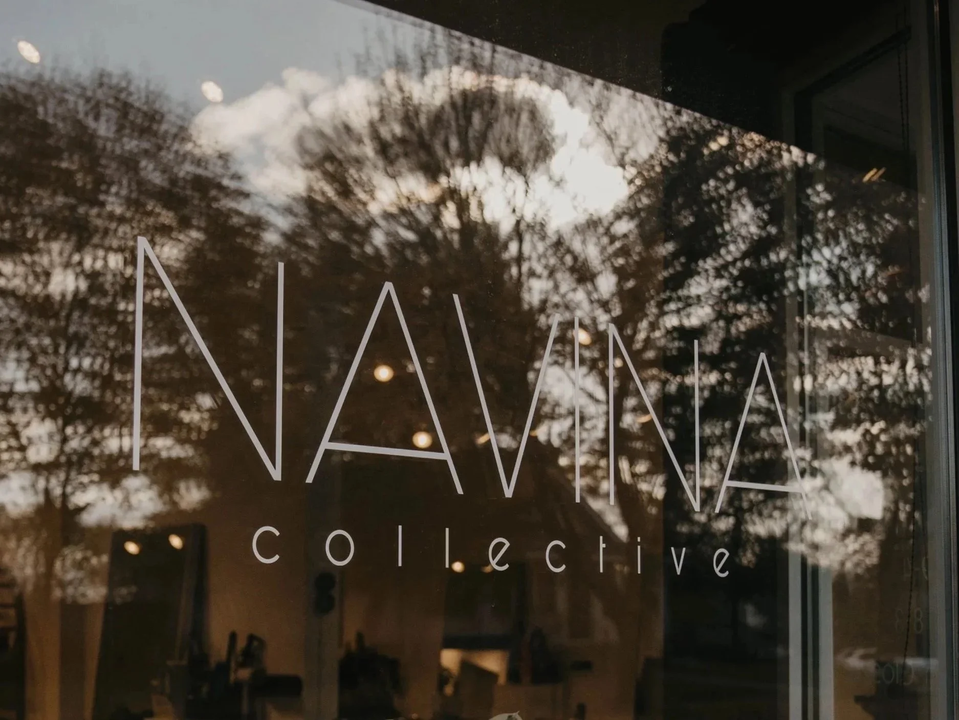 Reflection of trees and cloudy sky on glass window with text 'NAVina collective” Saint Paul, Minneapolis, twin cities salon in minnesota