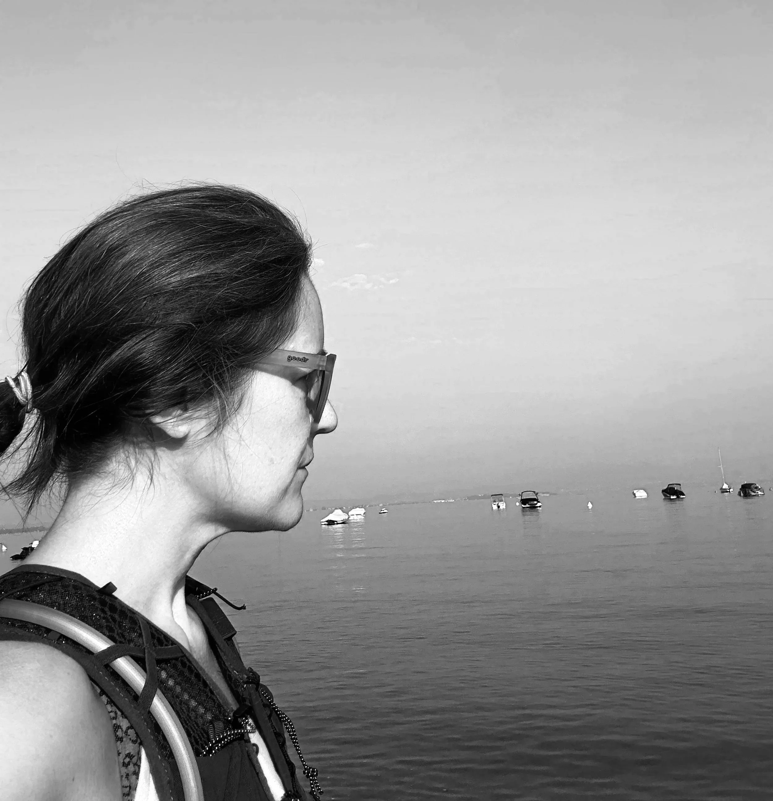 Black and white photo of a woman with glasses and a ponytail, looking out over a calm body of water with multiple boats anchored in the distance.