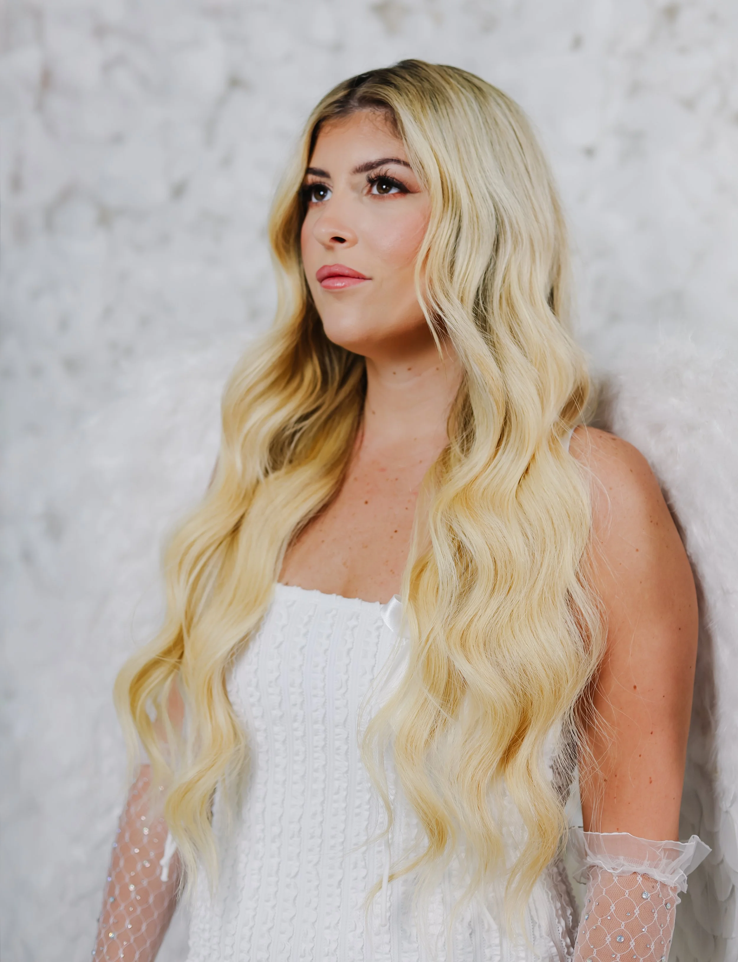 A woman with long, wavy blonde hair wearing a white dress stands against a white brick wall.