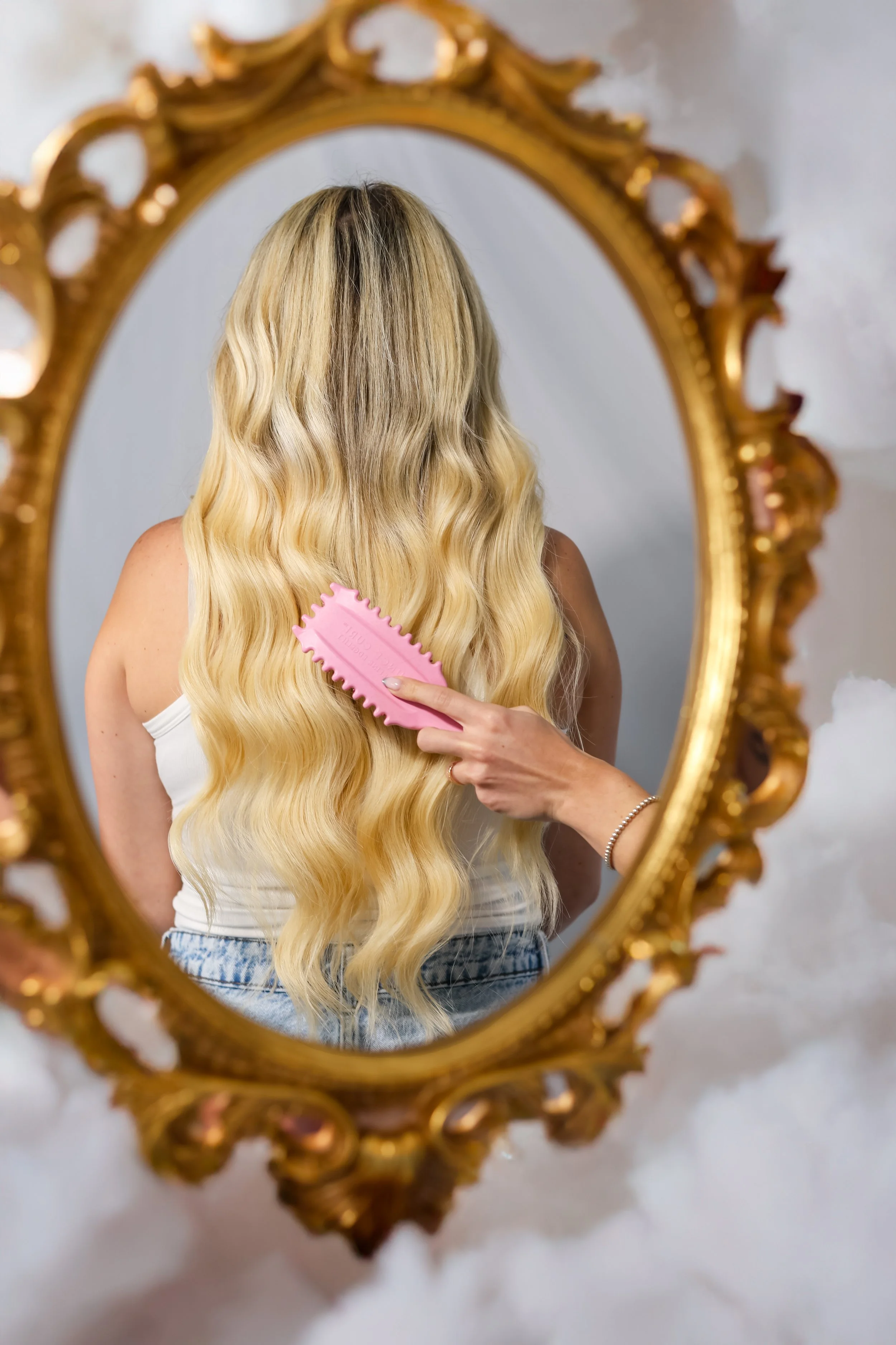A woman with long, wavy blonde hair brushing her hair in front of a decorative gold-framed mirror.