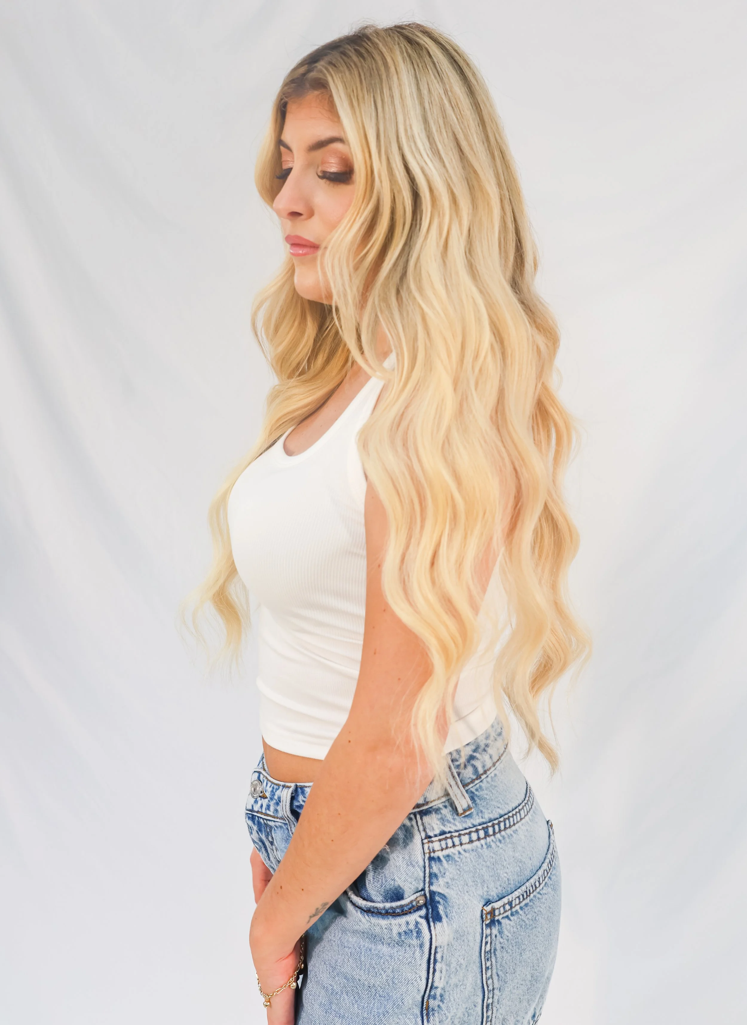 A woman with long, wavy blonde hair standing sideways against a plain white background, wearing a white sleeveless top and light blue jeans.