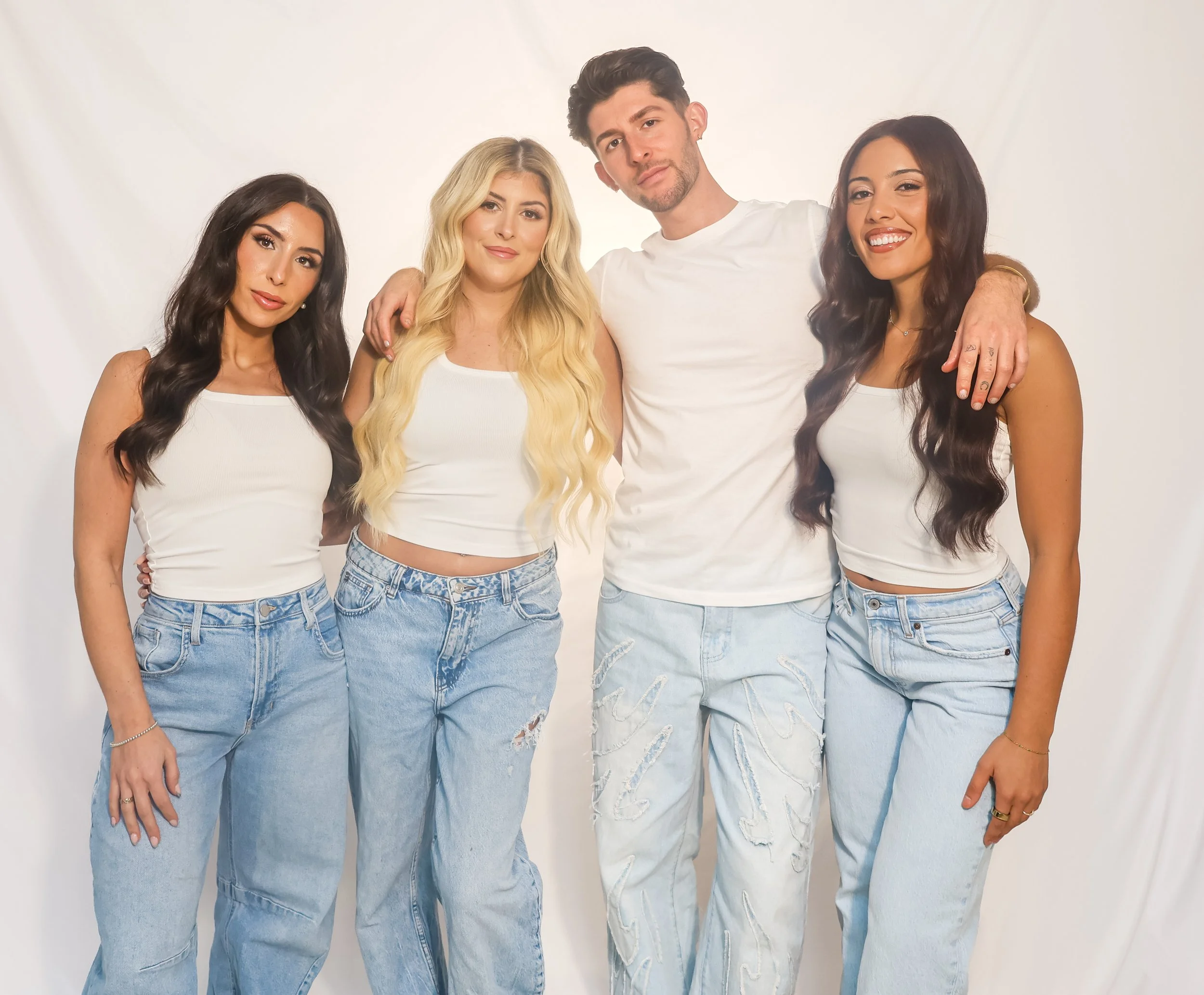 Four young adults standing together in front of a white backdrop, wearing casual white tops and light blue jeans, smiling and posing for the camera.