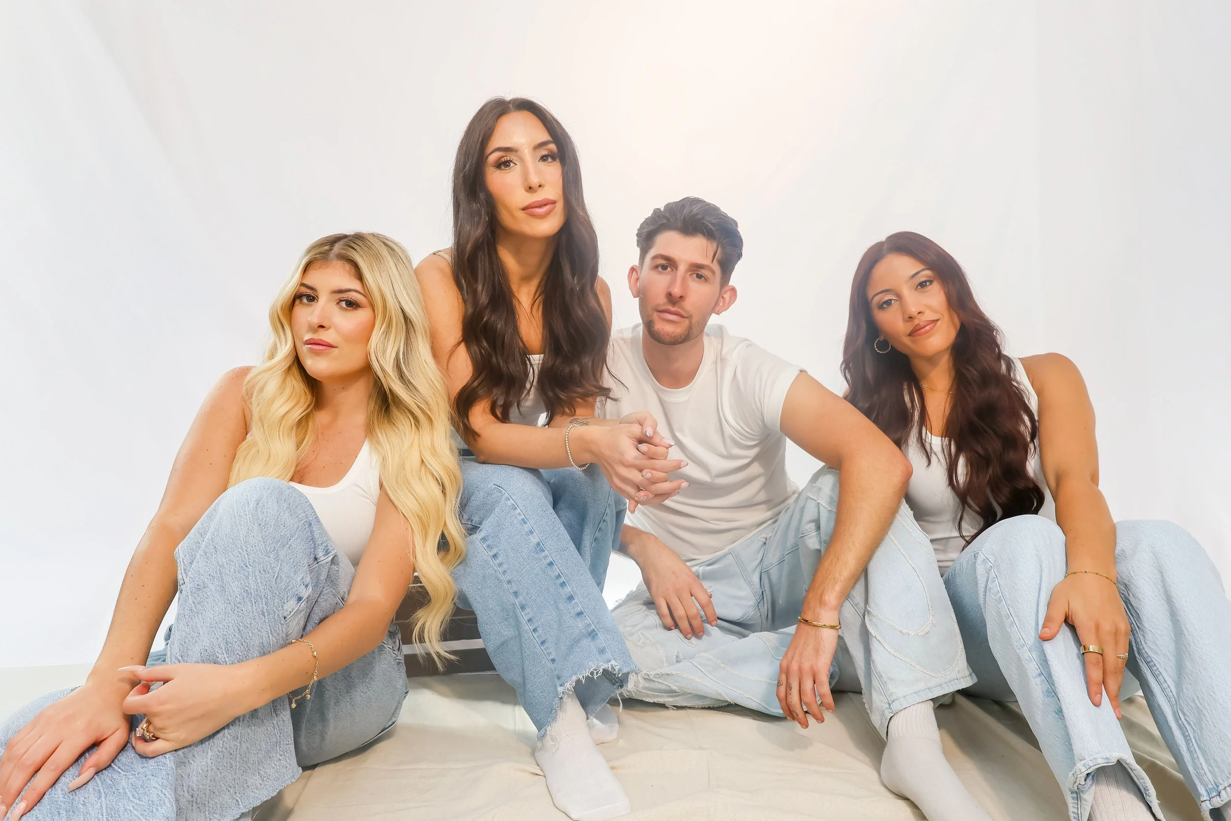 A group of four young adults sitting together on a bed against a plain white background, casually dressed in light-colored clothing.