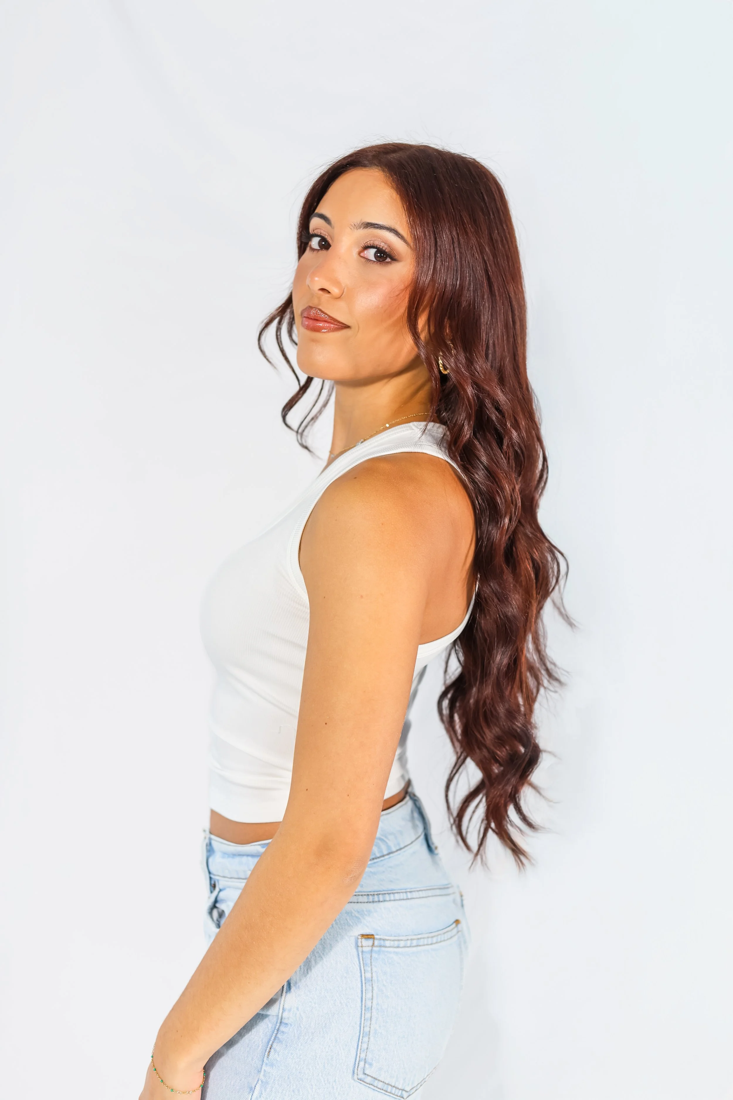 A woman with long wavy reddish-brown hair looking to the side, wearing a white sleeveless top and light blue jeans, standing against a plain white background.