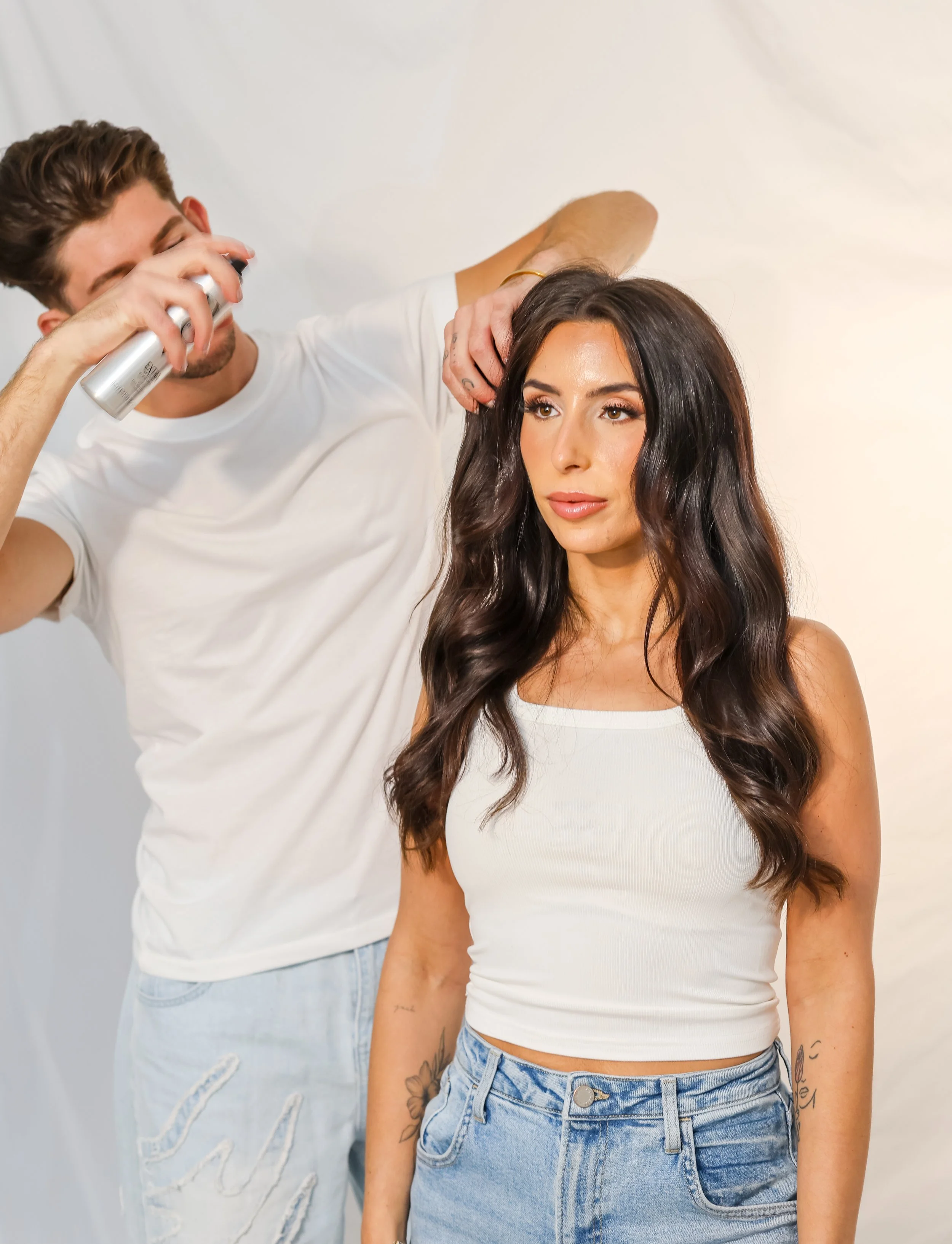 A woman with long dark hair in a white ribbed tank top and blue jeans has her hair styled by a hairstylist using hairspray.