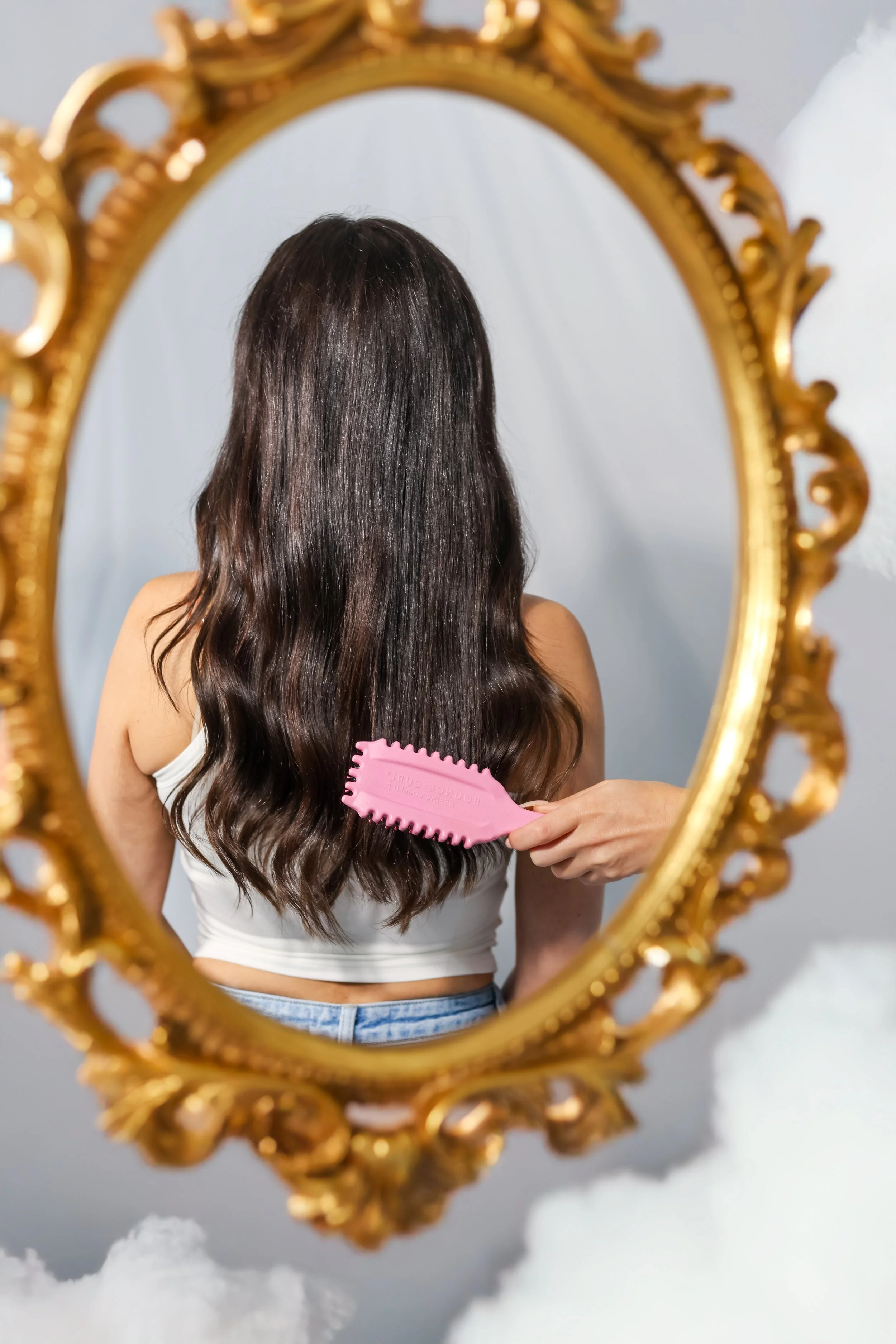 A woman with long, wavy brown hair is brushing her back hair in front of an ornate gold mirror, with a cloud-like decoration at the bottom.