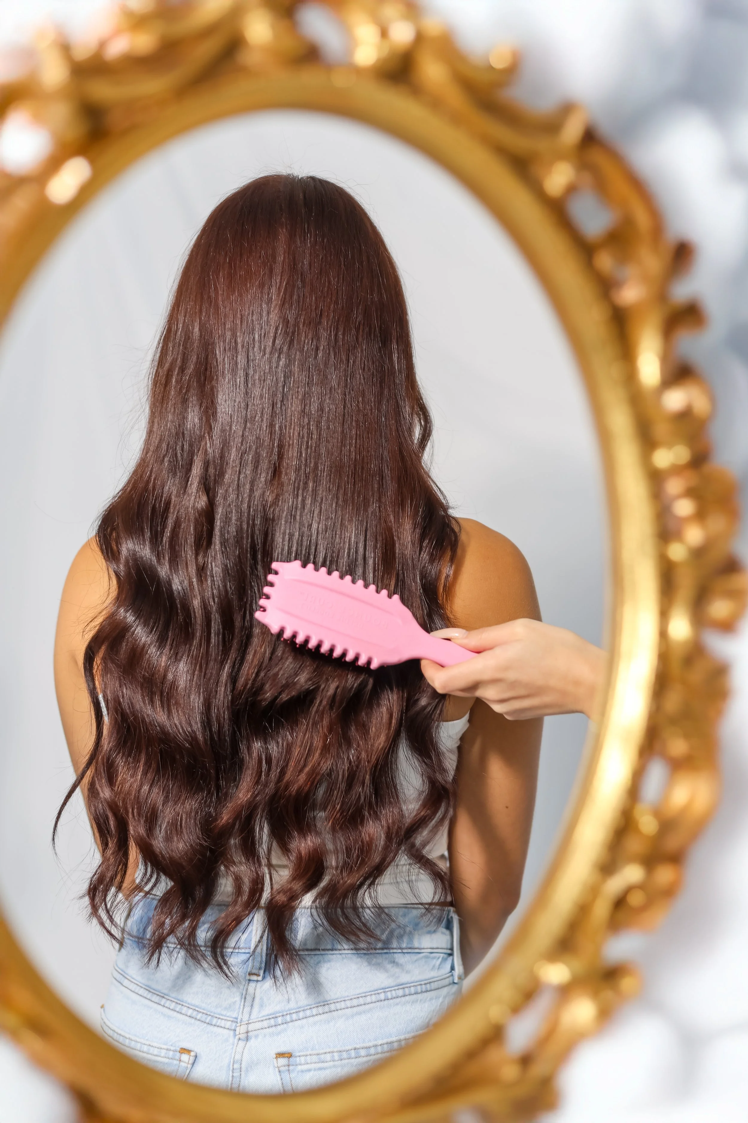 A person with long, wavy brown hair is brushing their hair while looking into a decorative gold-framed mirror.