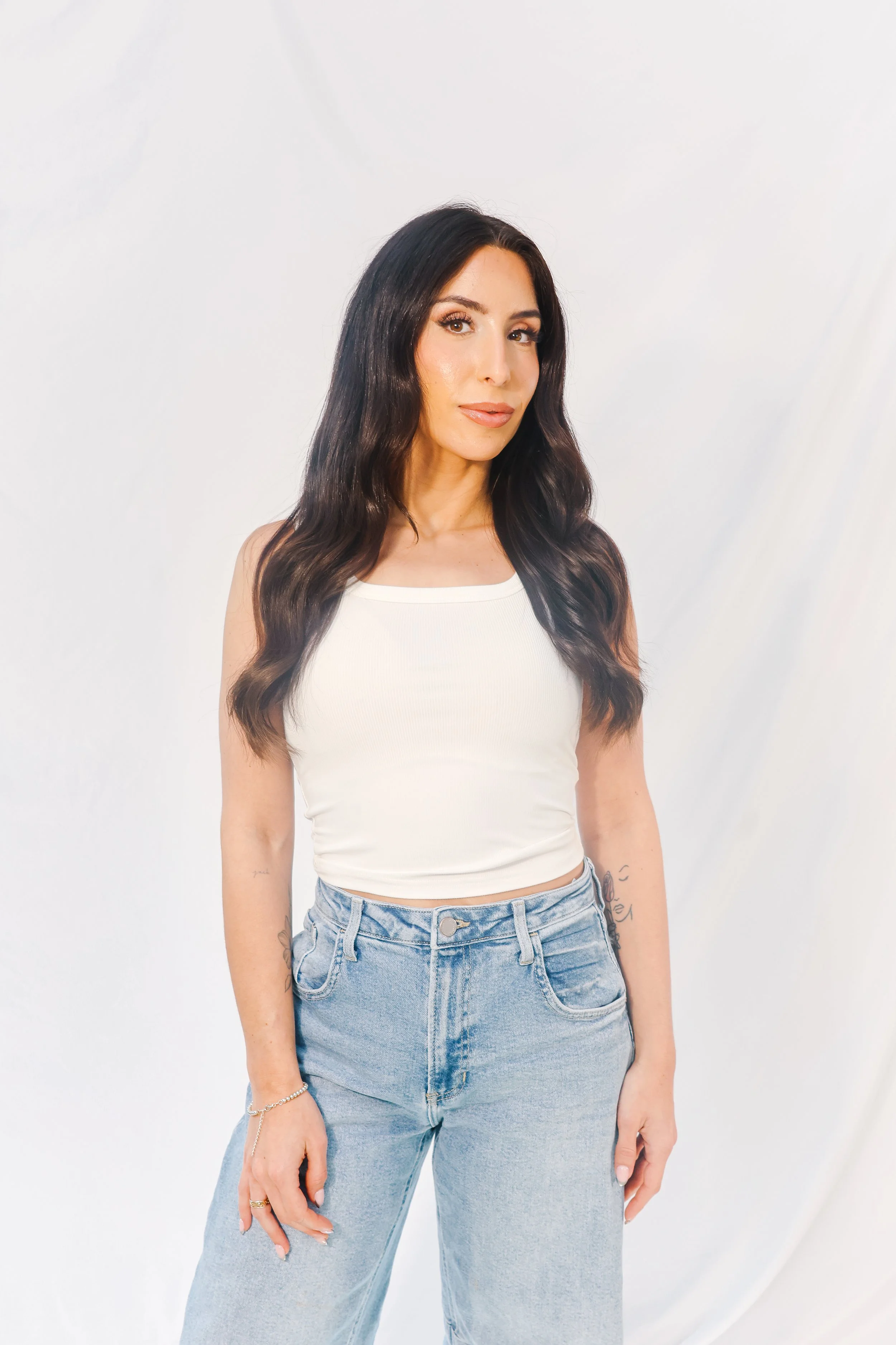 A woman with long dark hair, wearing a white sleeveless top and light blue jeans, standing in front of a plain white background.