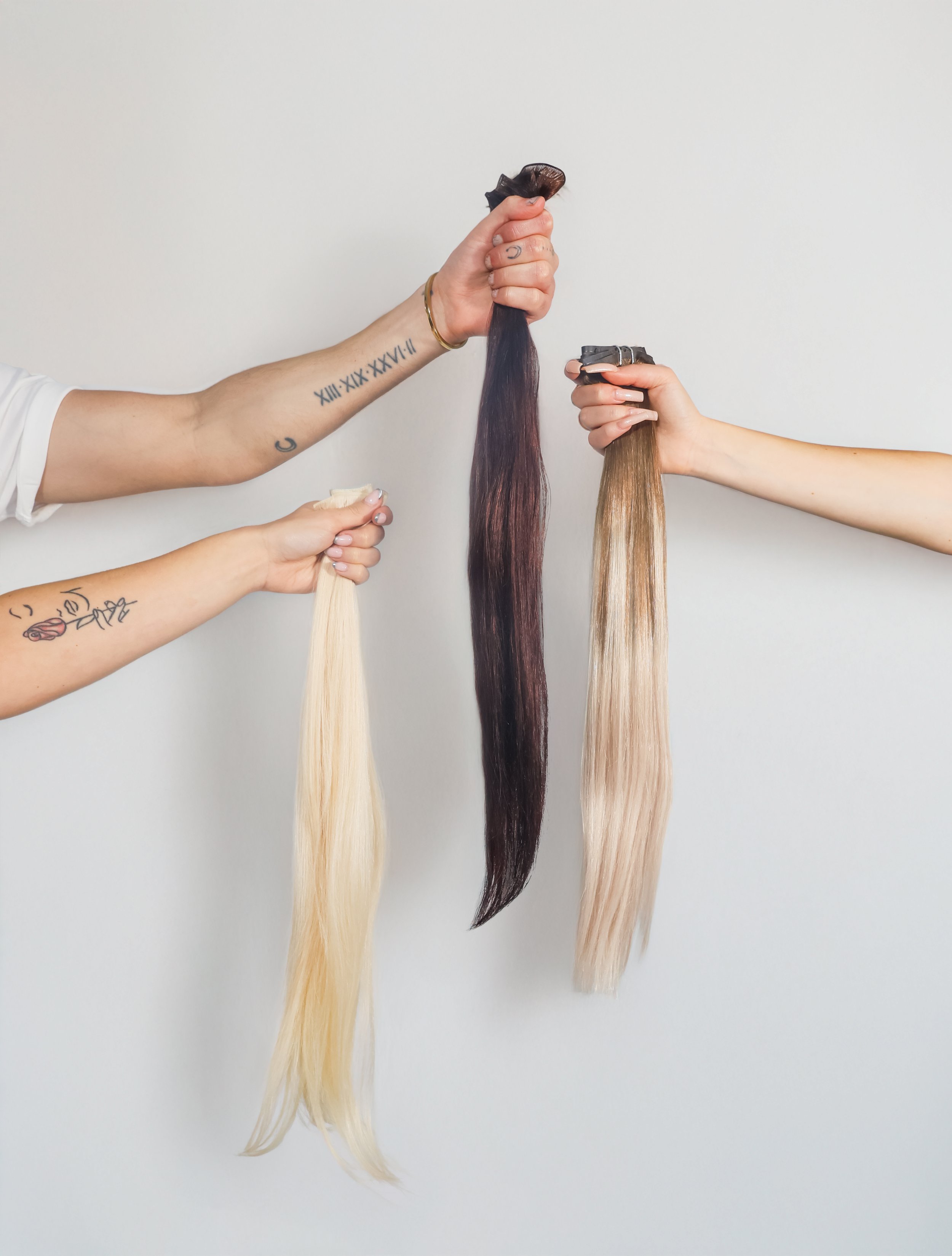 Three hands holding hair extensions of different colors against a plain white background.