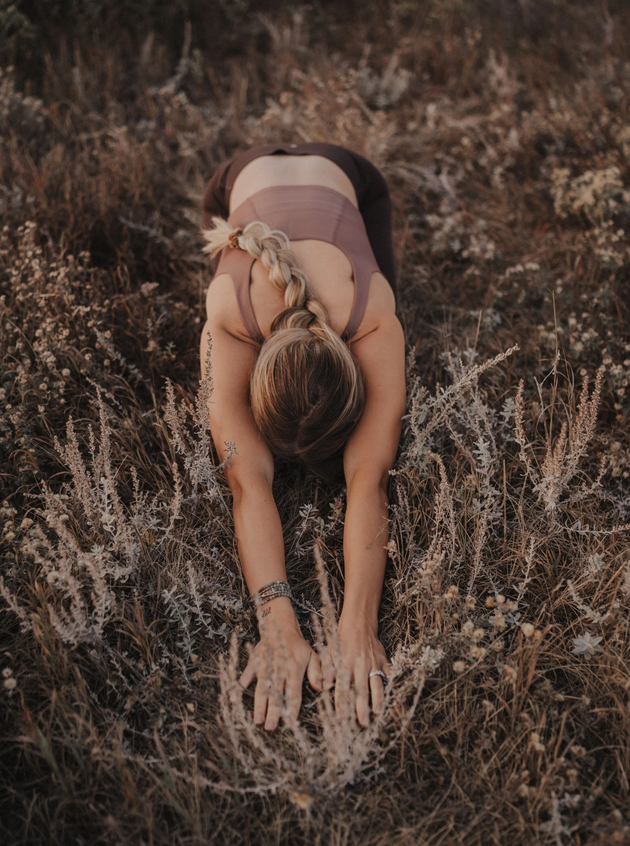 A woman with long blonde hair in a braid practicing yoga outdoors on dry grass, stretching forward with her arms extended and hands on the ground.