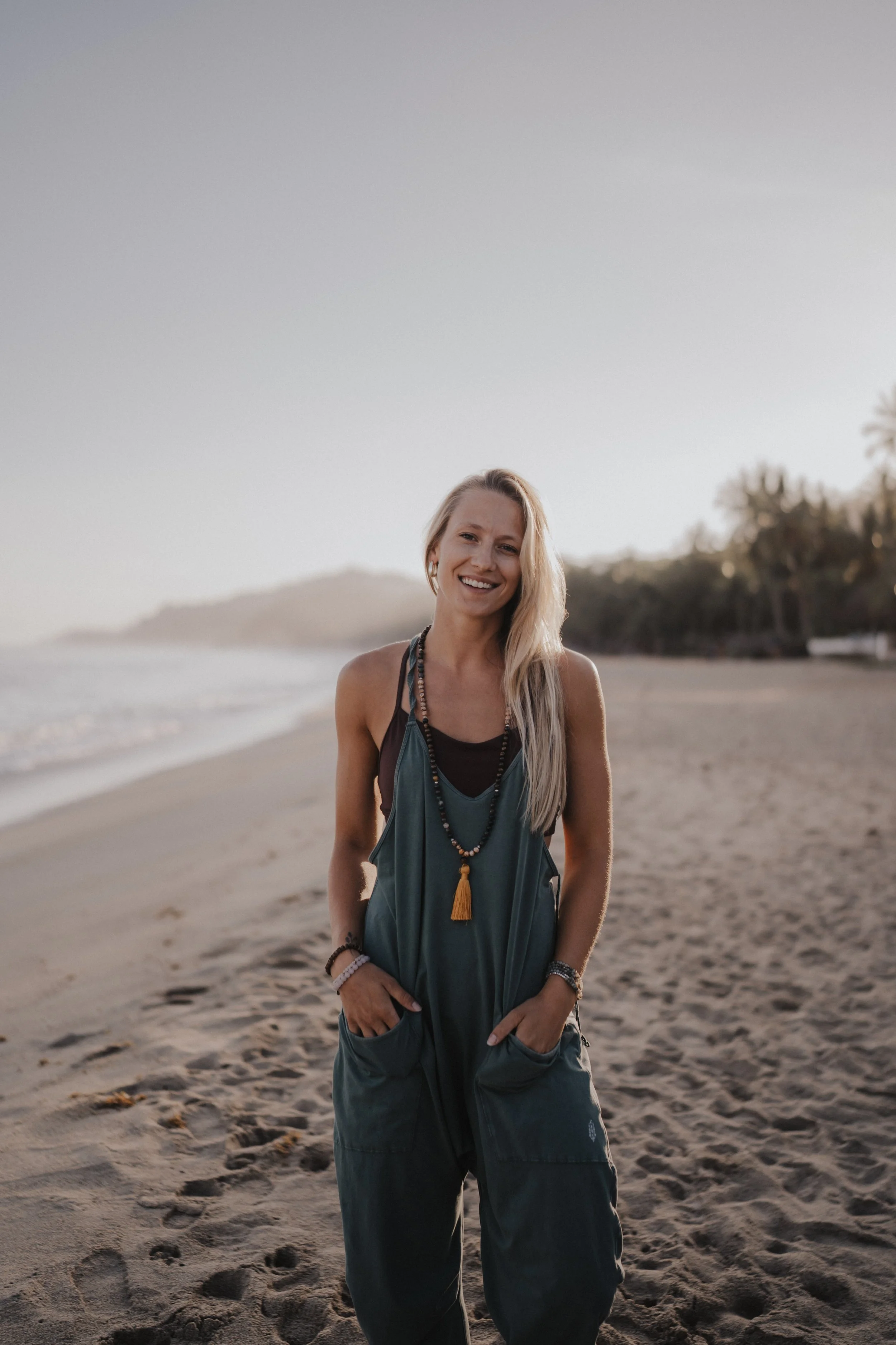 Young woman standing on a sandy beach smiling with the ocean and trees in the background during sunset.