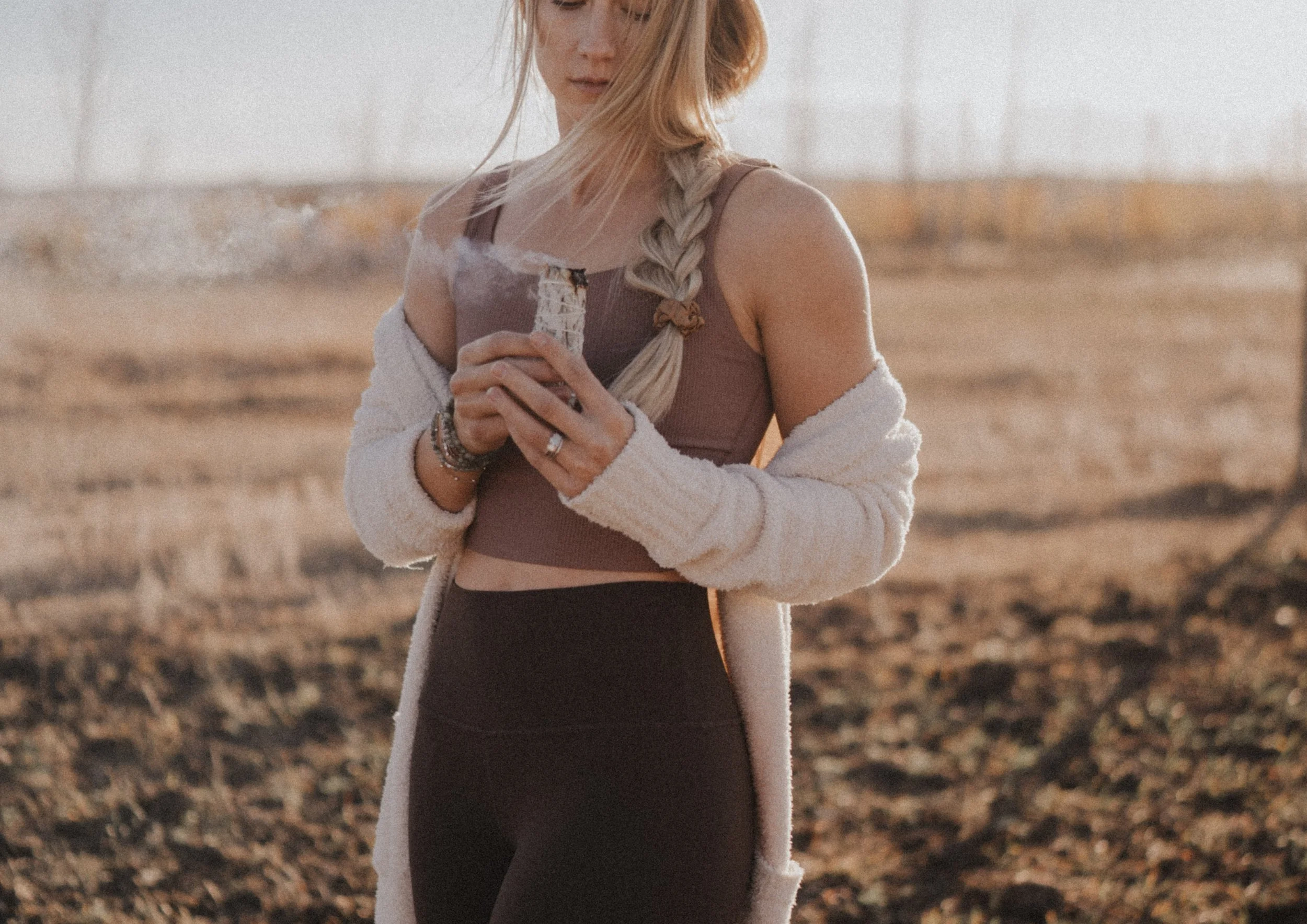 A woman with long blonde hair in a braid, wearing a brown crop top, black leggings, and a white cardigan, holding a steaming mug outdoors during sunset.