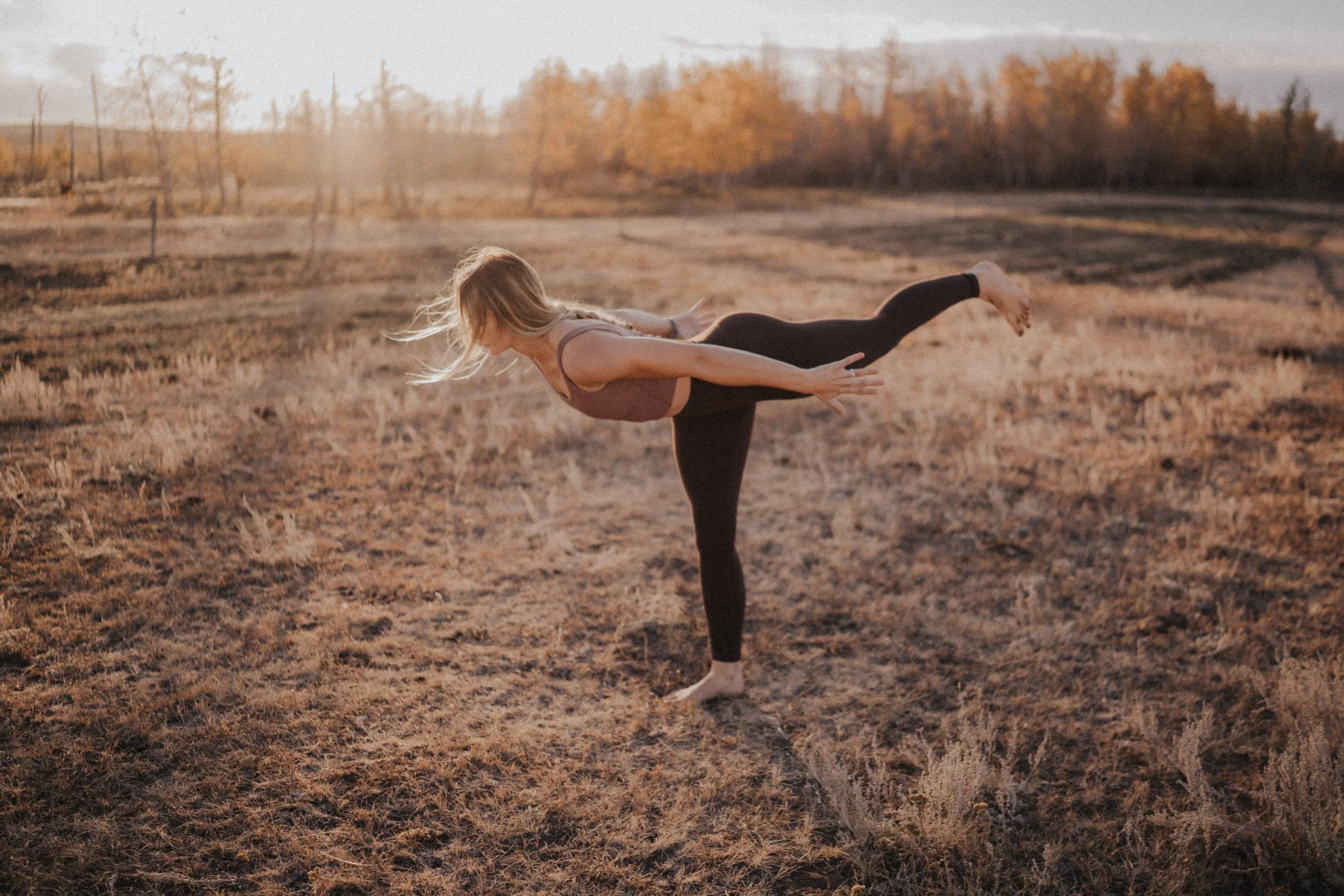 Nicole, owner of Sage the Sol, practicing yoga outdoors in a field during sunset, in Warrior III Pose.
