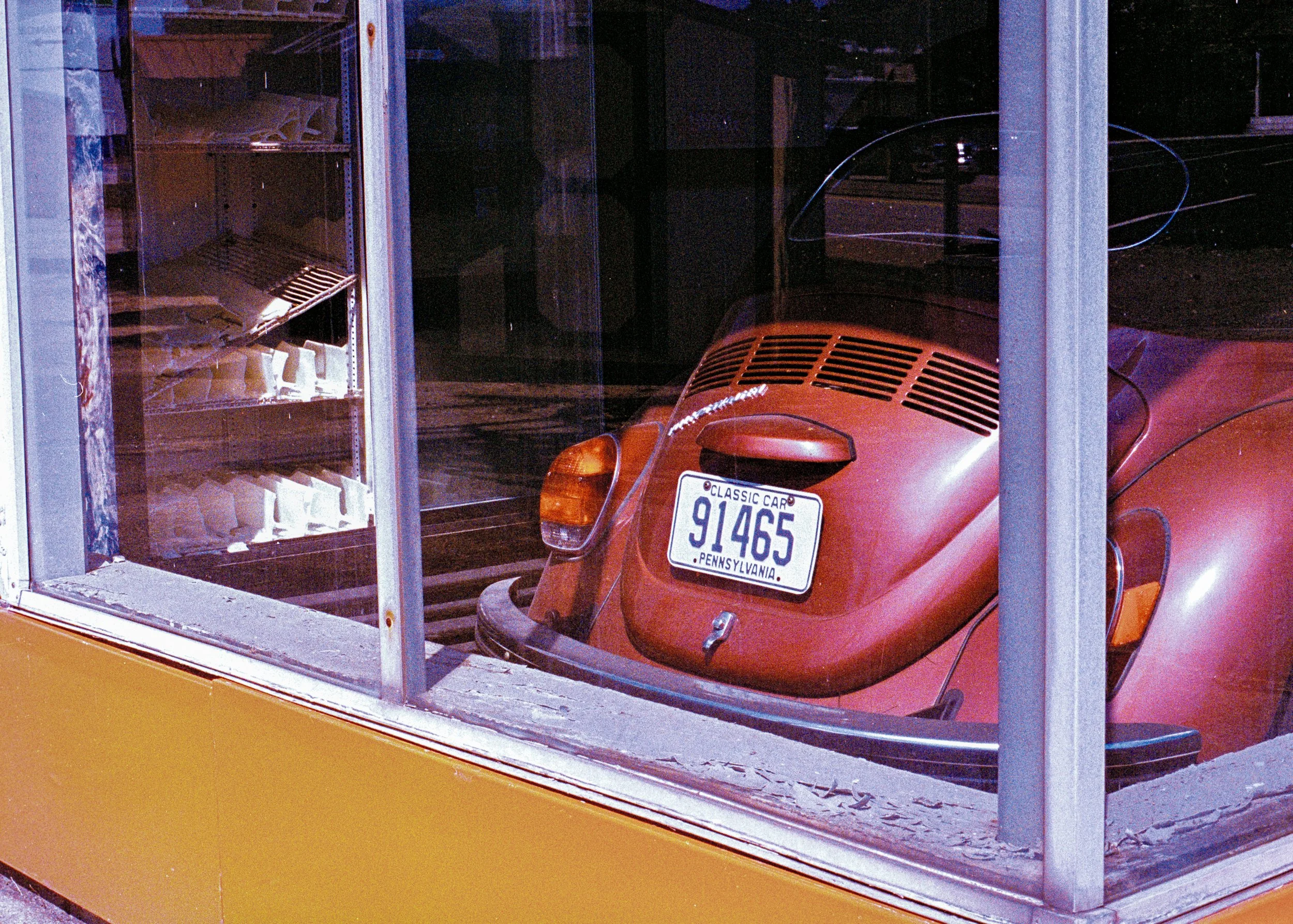 A red vintage Volkswagen Beetle parked inside a glass display case, with a Pennsylvania license plate reading 91465, labeled 'Classic Car.' A bakery display with baked goods is visible on the left side behind the glass.