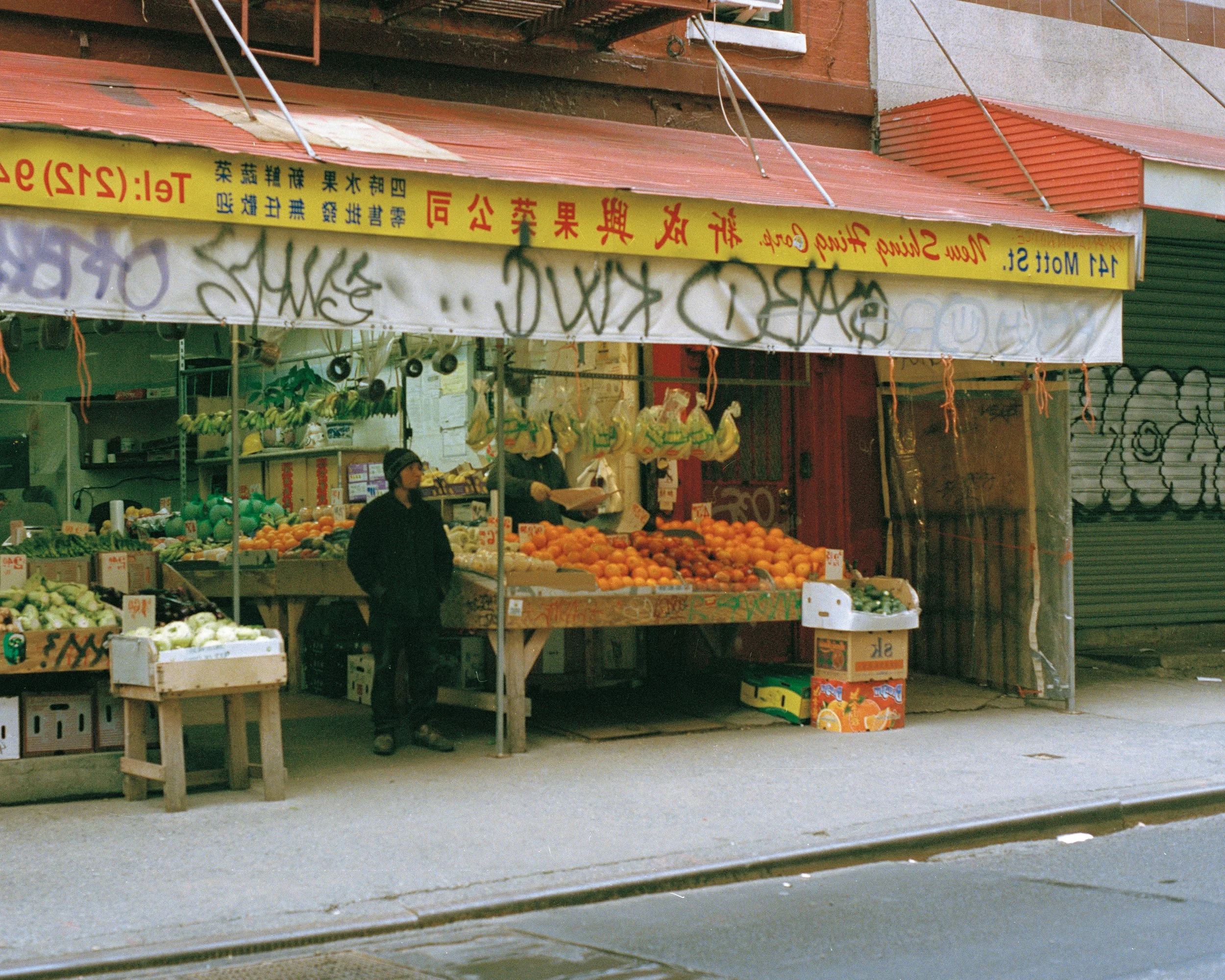 A street fruit stand in an urban area with various fresh fruits, a person in black clothing and a hat, and graffiti-covered walls in the background.