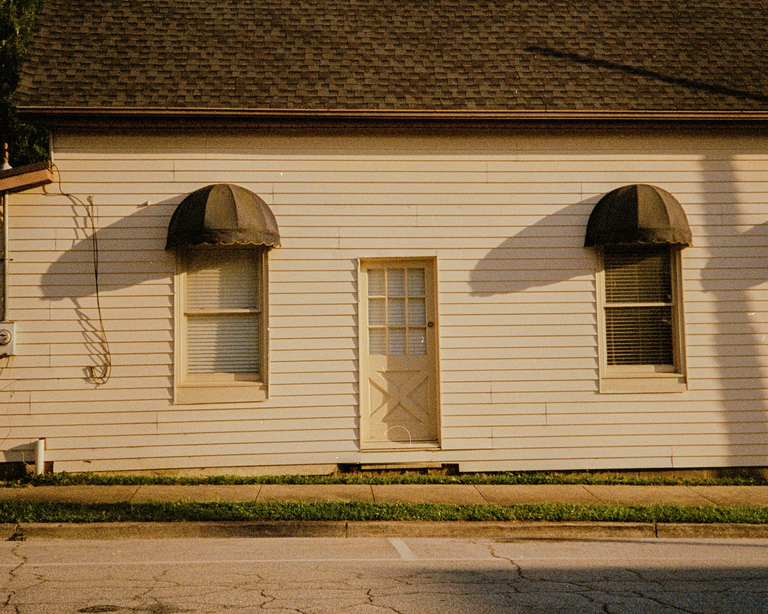 Front of a small, white, wooden house with two windows and an exterior door in the middle. Each window has a black, semi-circular awning casting shadows on the wall. The house has a shingled roof and is next to a sidewalk and street.