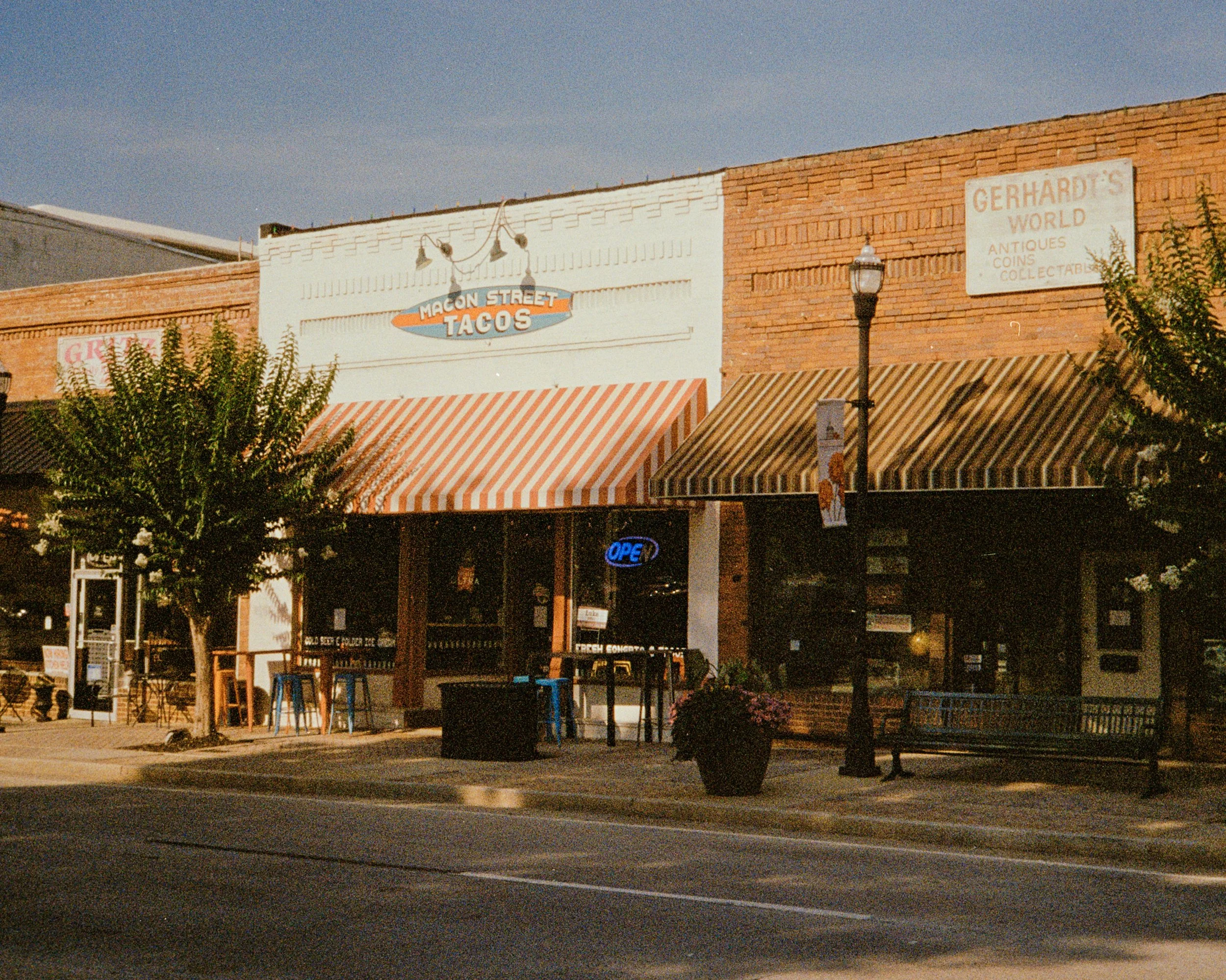 A small town storefront with a sign reading "Macon Street Tacos," striped awnings, a lit "Open" sign, and outdoor seating along the sidewalk. There are trees, a streetlamp, and vintage-style buildings in the background.