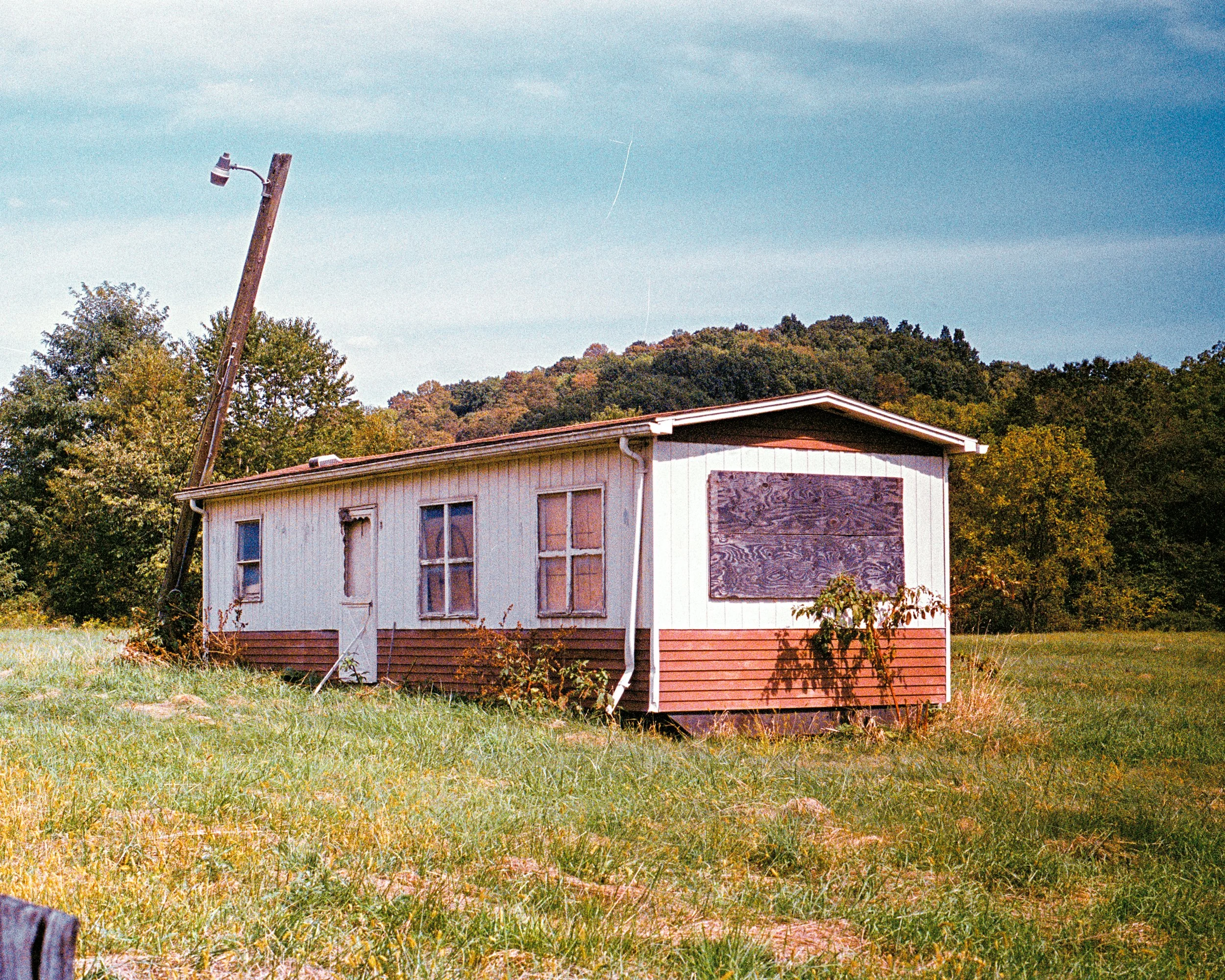 An abandoned house with broken windows and boarded-up sections, situated in a grassy field with trees and hills in the background, and a leaning utility pole to the left.