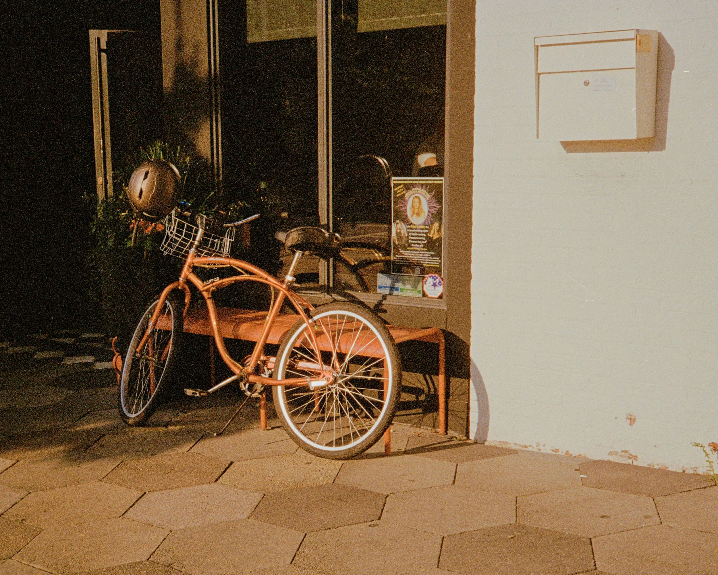 A peach-colored bicycle with a basket and helmet parked outside a building at night, with a poster in the window and a white wall with a mailbox.