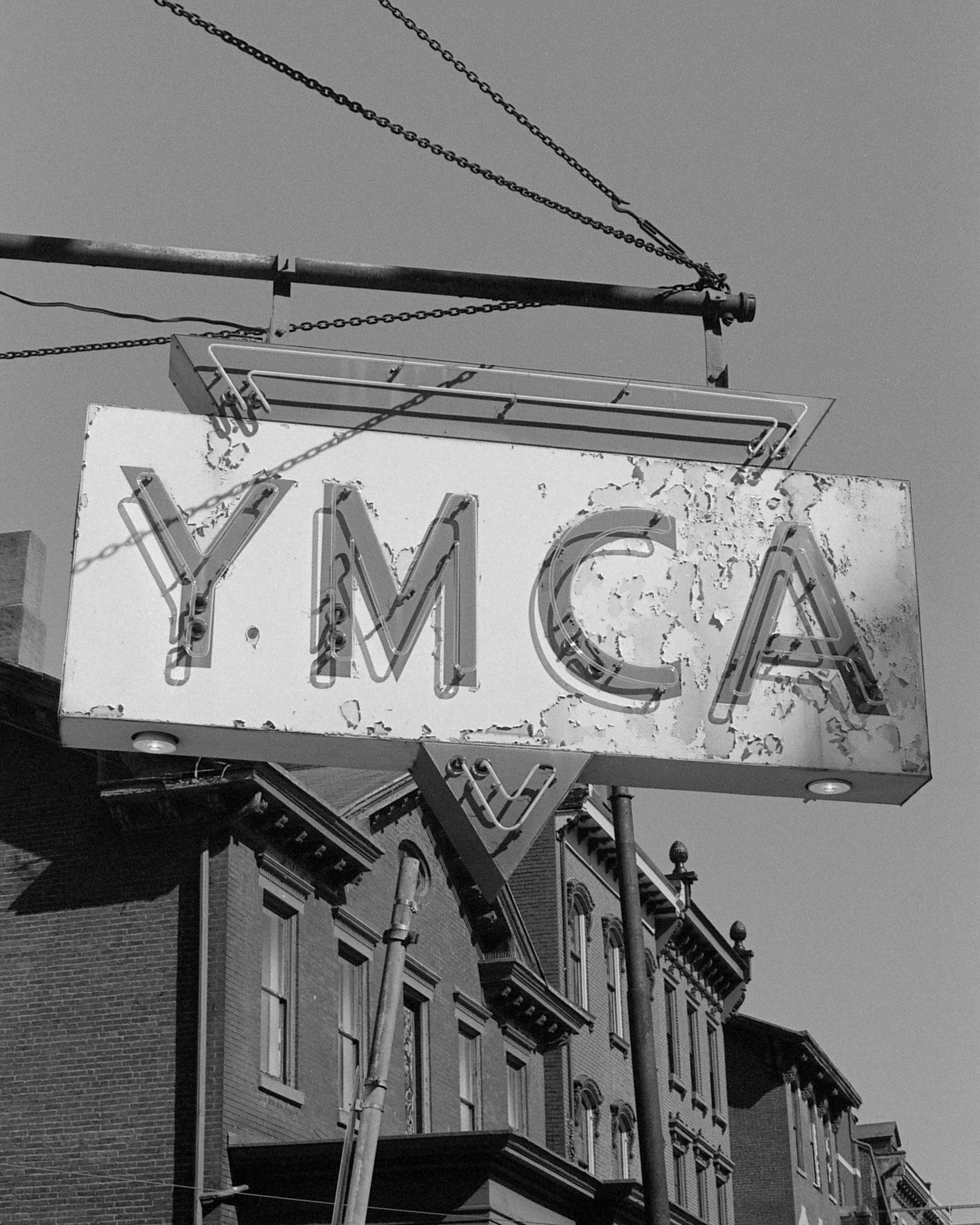 A weathered neon sign with the letters 'Y.M.C.A' hanging on a street corner. The sign appears old and peeling, with buildings and power lines visible in the background.