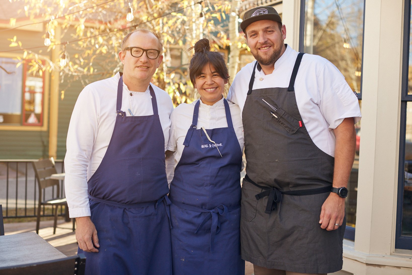 Three chefs in white shirts and aprons smiling outside, standing close together with trees and a building in the background.