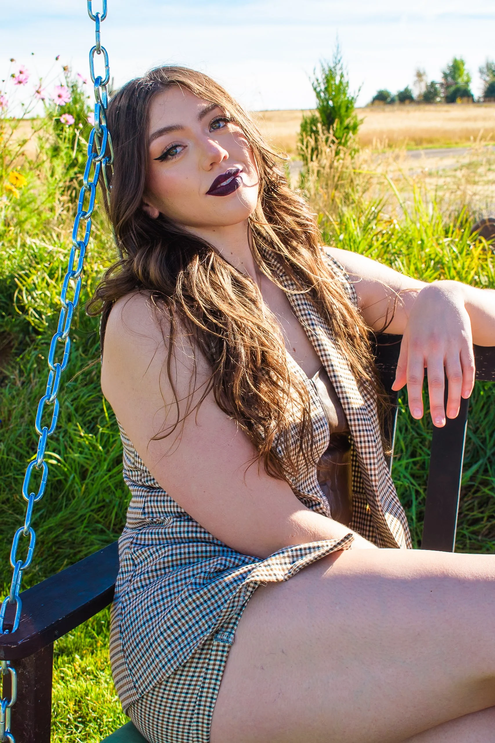 A young woman with wavy brown hair and dark lipstick sits on a swing outdoors, surrounded by greenery and meadow, wearing a sleeveless checkered dress.