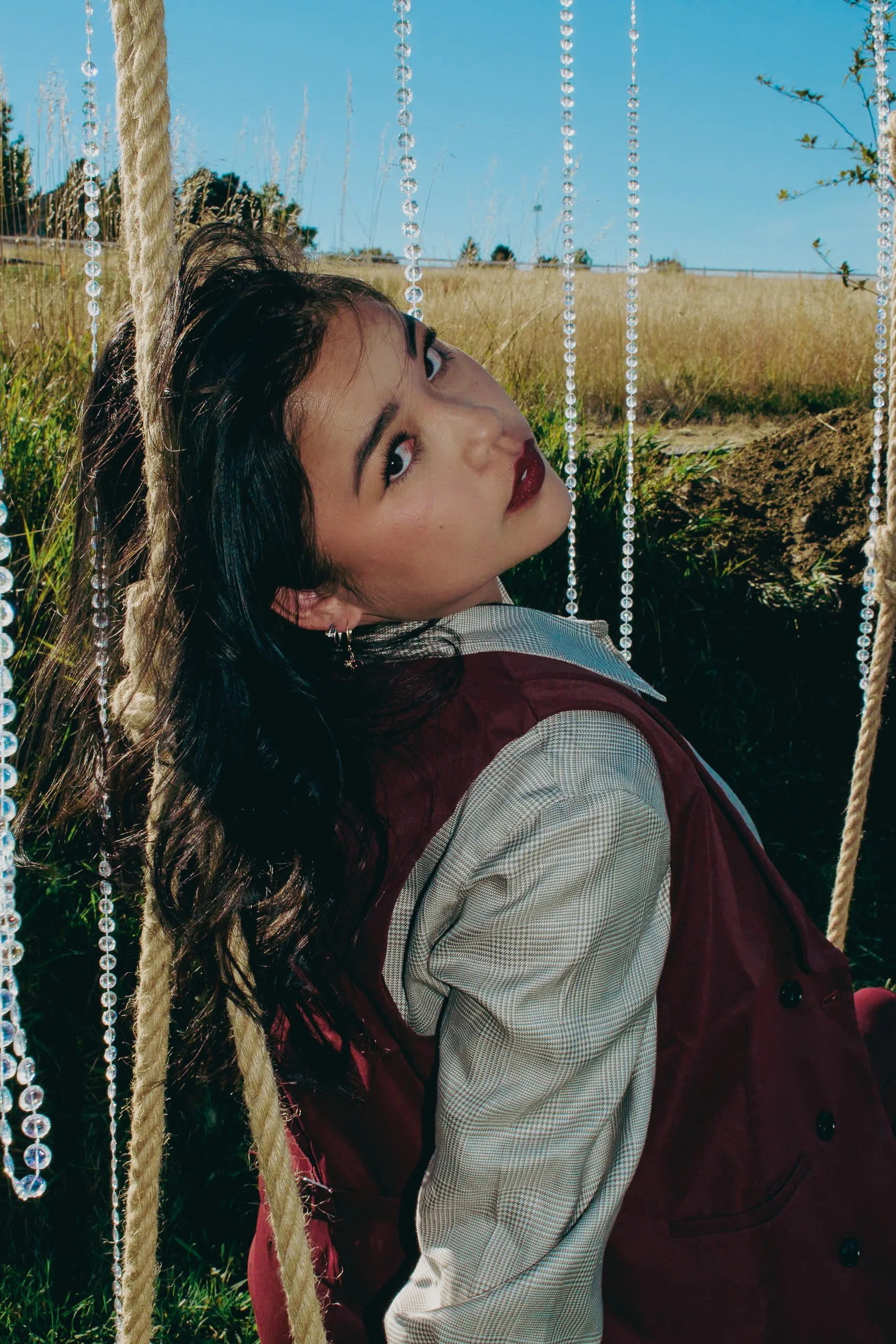 A woman with dark hair and makeup leaning against a swinging seat outdoors, with grass, bushes, and a blue sky in the background, decorated with hanging beads or crystals.