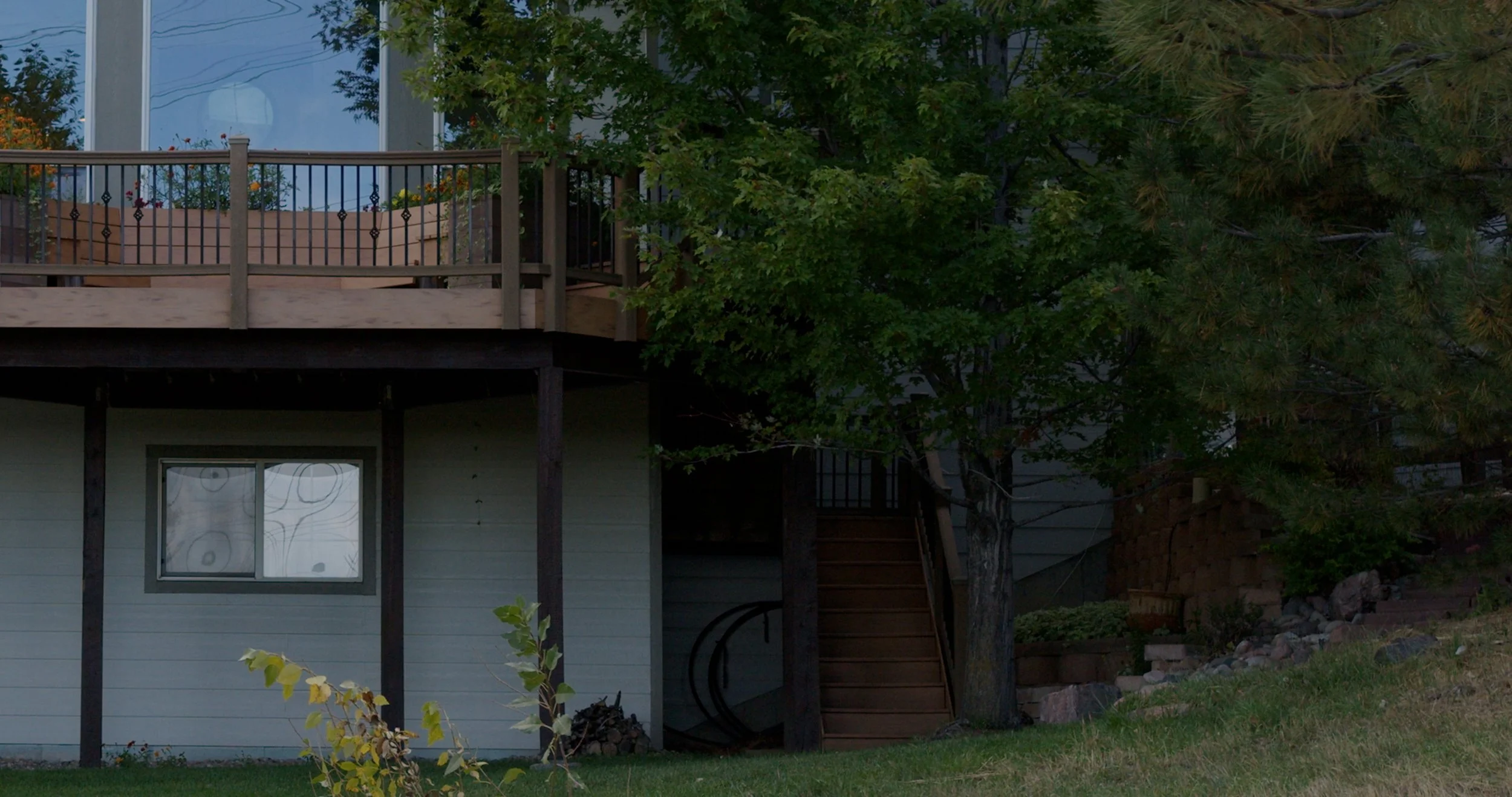 A house with a small window on the lower level, a staircase on the side leading to a balcony with flower pots, a large tree beside the house, and a grassy yard with rocks.