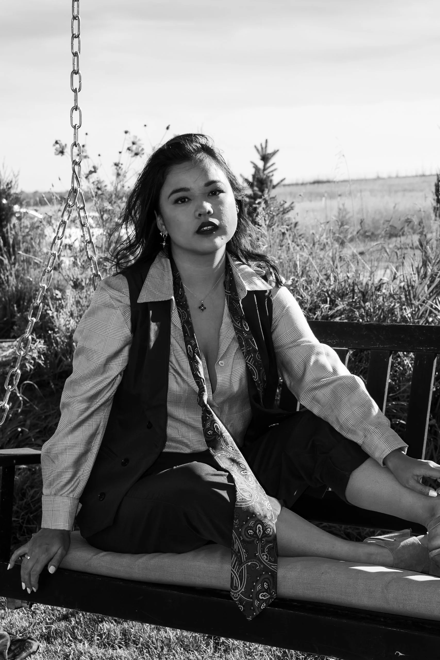 A young woman with dark hair and dark lipstick sitting on a swing outdoors in a field. She is wearing a checked shirt, a vest, and a patterned scarf, with an open-leg pose and a serious expression.