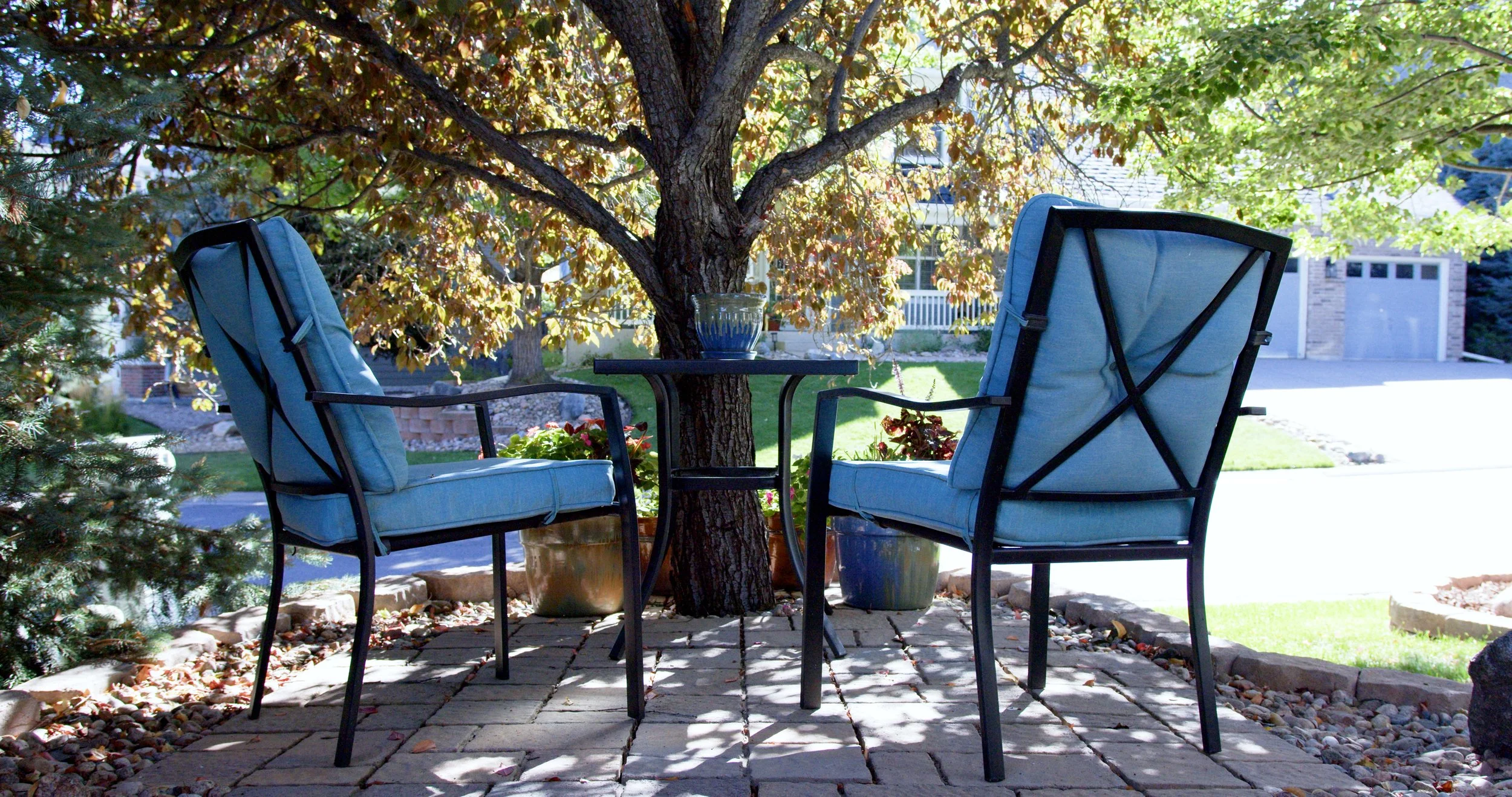 Two blue cushioned patio chairs facing each other with a small round black table in between, set under a leafy tree in a backyard, with a house and garage in the background.