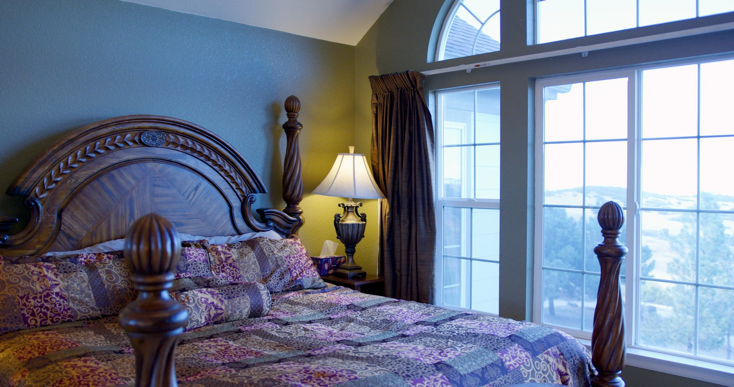 A bedroom with a wooden four-poster bed, purple and gold patterned bedding, a bedside lamp, and large windows showing a daytime outdoor view.