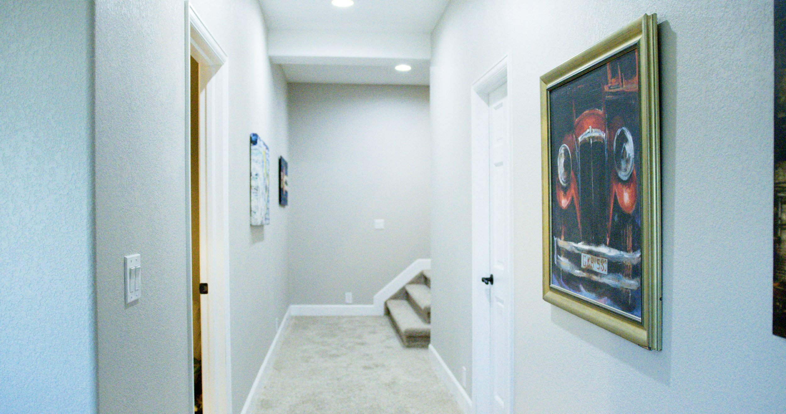 Hallway with light-colored walls, carpeted stairs, and framed artwork including a painting of a red vintage car on the right wall.