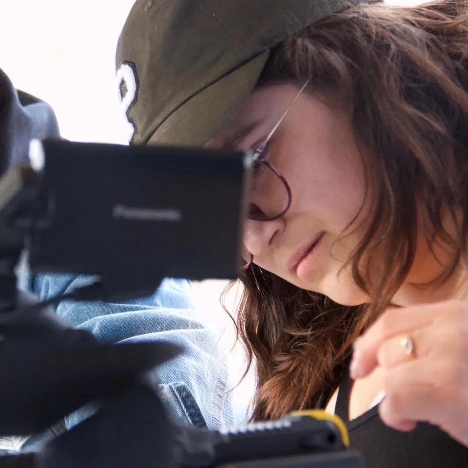 A woman with glasses and curly hair wearing a baseball cap, looking down at a camera or device.