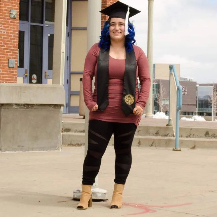 A young woman with blue hair wearing a graduation cap, gown sash, and black leggings standing outside in front of steps, smiling.