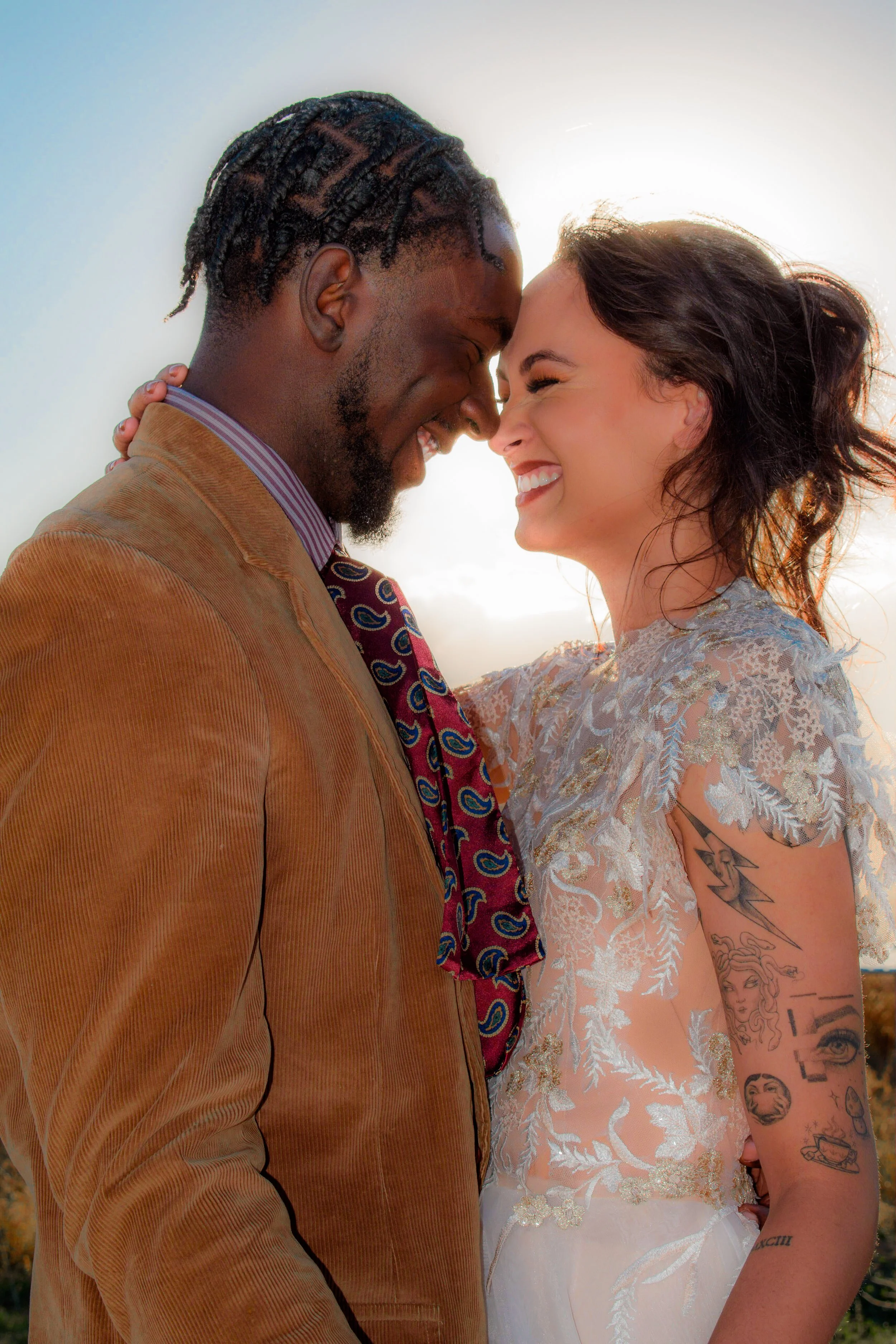 A smiling couple with foreheads touching, outdoors, backlit by the sun, in a close embrace.