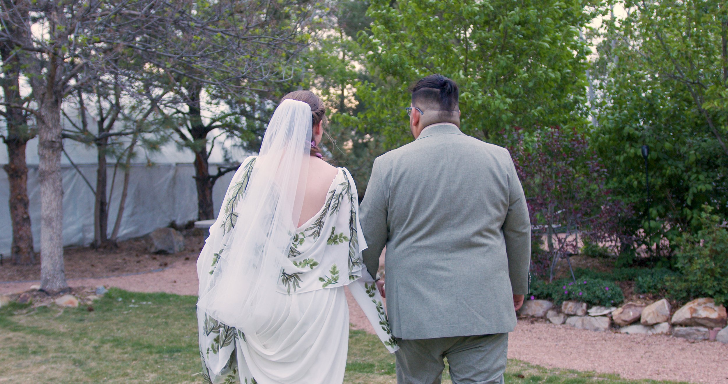 A bride and groom walking outdoors on a garden path, surrounded by trees and greenery.