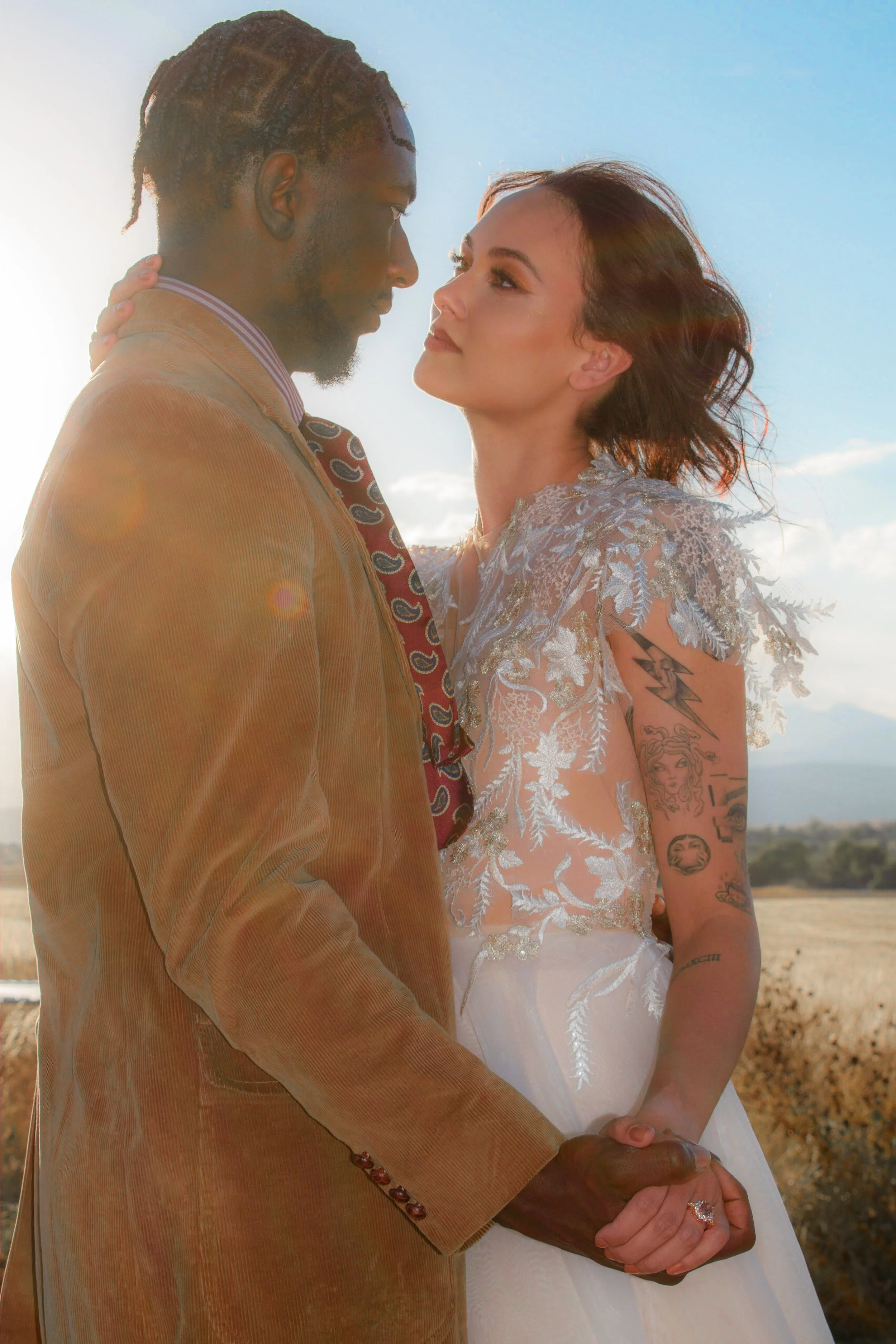 A couple holding hands and looking into each other's eyes outdoors on a sunny day. The man is wearing a brown suit and the woman is wearing a lacy, embroidered dress with tattoos on her arm.