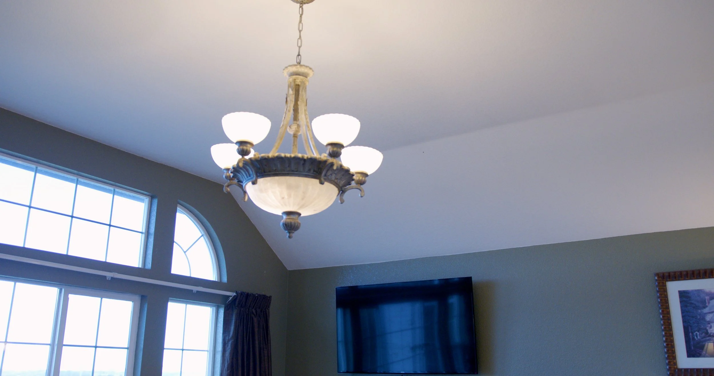 Ceiling chandelier with multiple white glass globes hanging from a chain in a living room with a flat-screen TV, a window with a semi-circular top, and a framed picture on the wall.