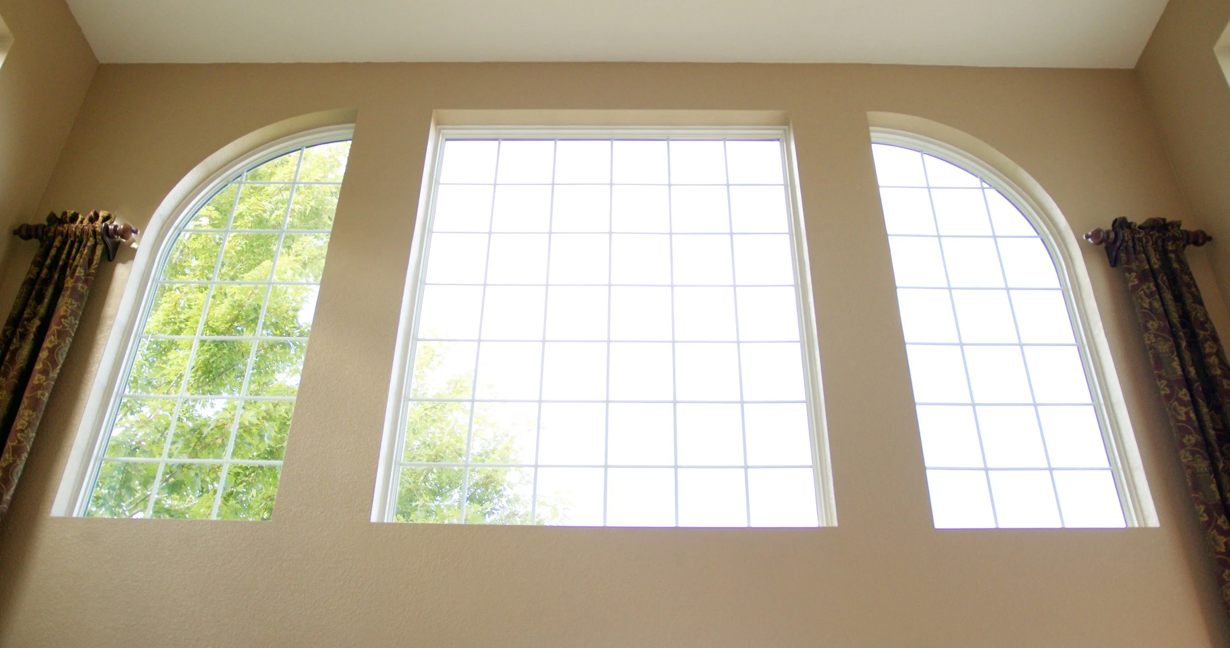 Three large windows with grid panes, two arched and one rectangular, in a beige wall with curtains on either side, overlooking green trees outside.