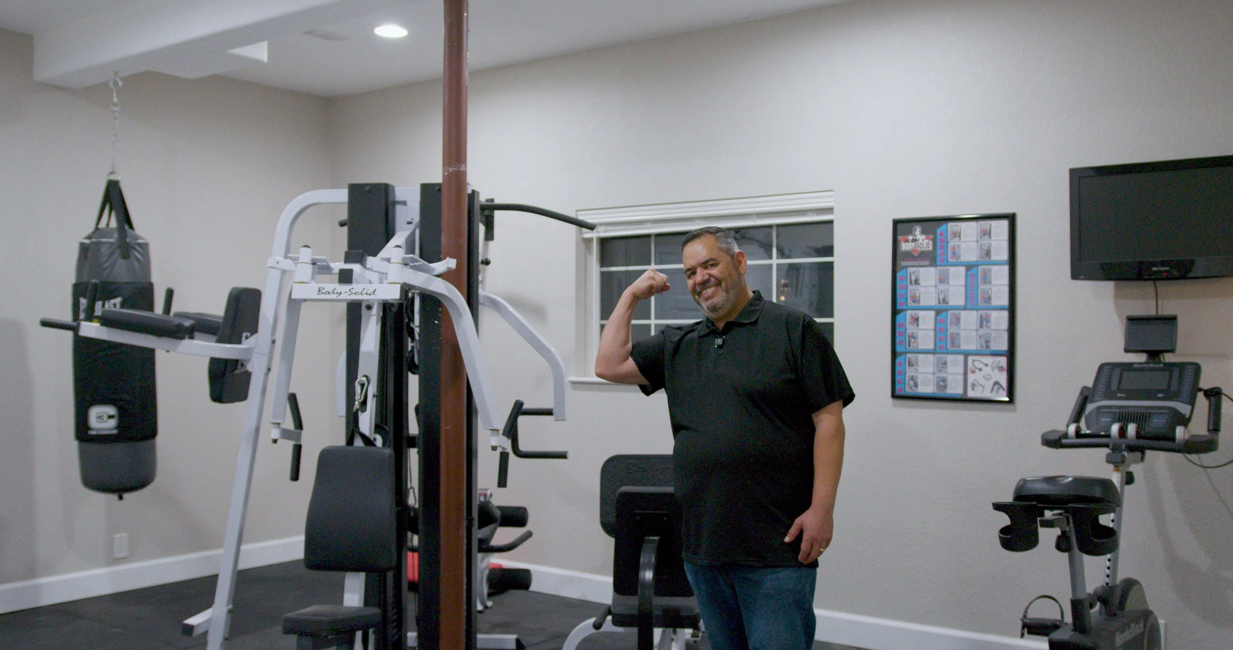 A man in a black shirt and jeans smiling and flexing his right arm in a home gym with exercise equipment like a punching bag, weight machine, and stationary bike.