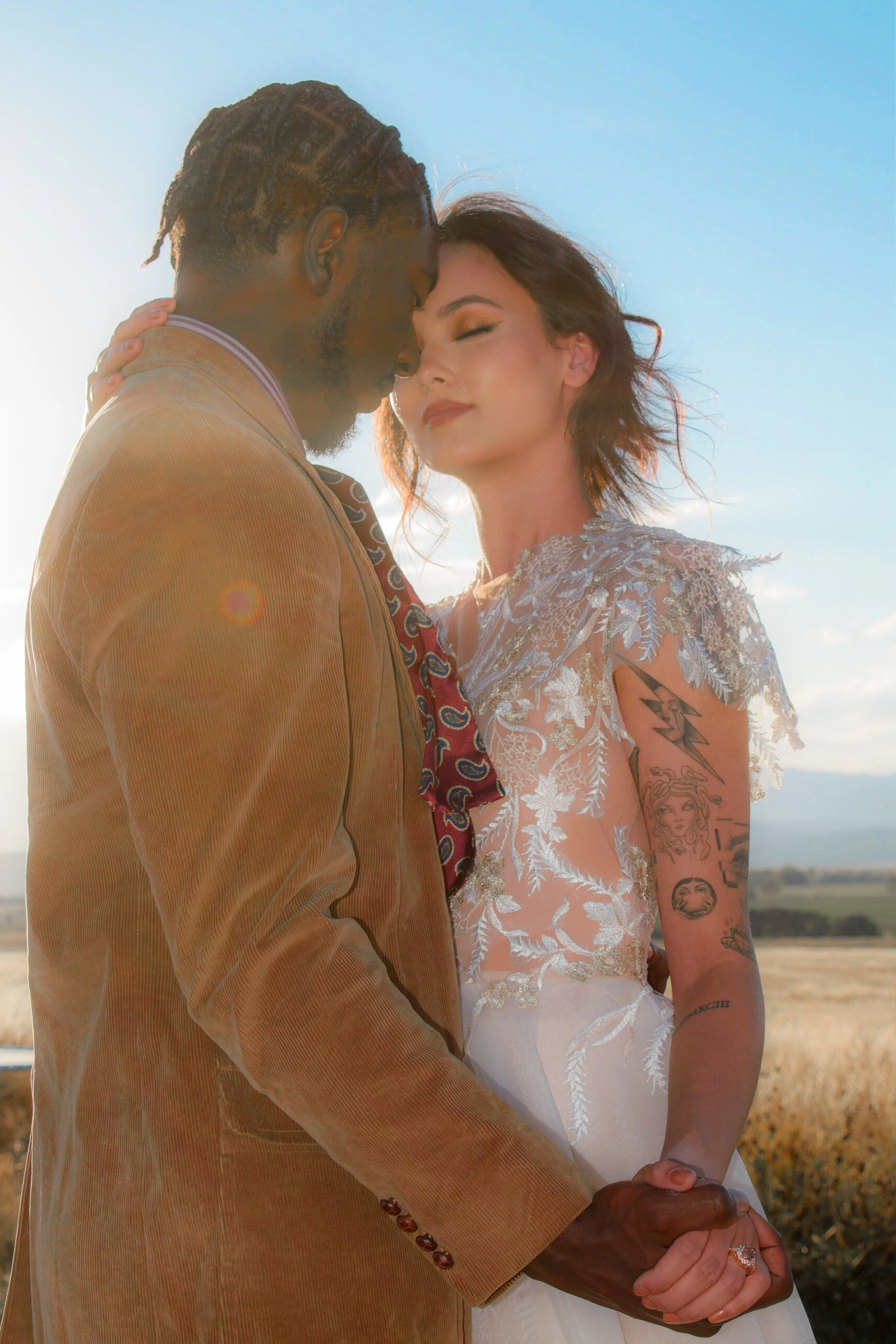 A couple in wedding attire holding hands and embracing outdoors with a rural landscape and blue sky in the background.