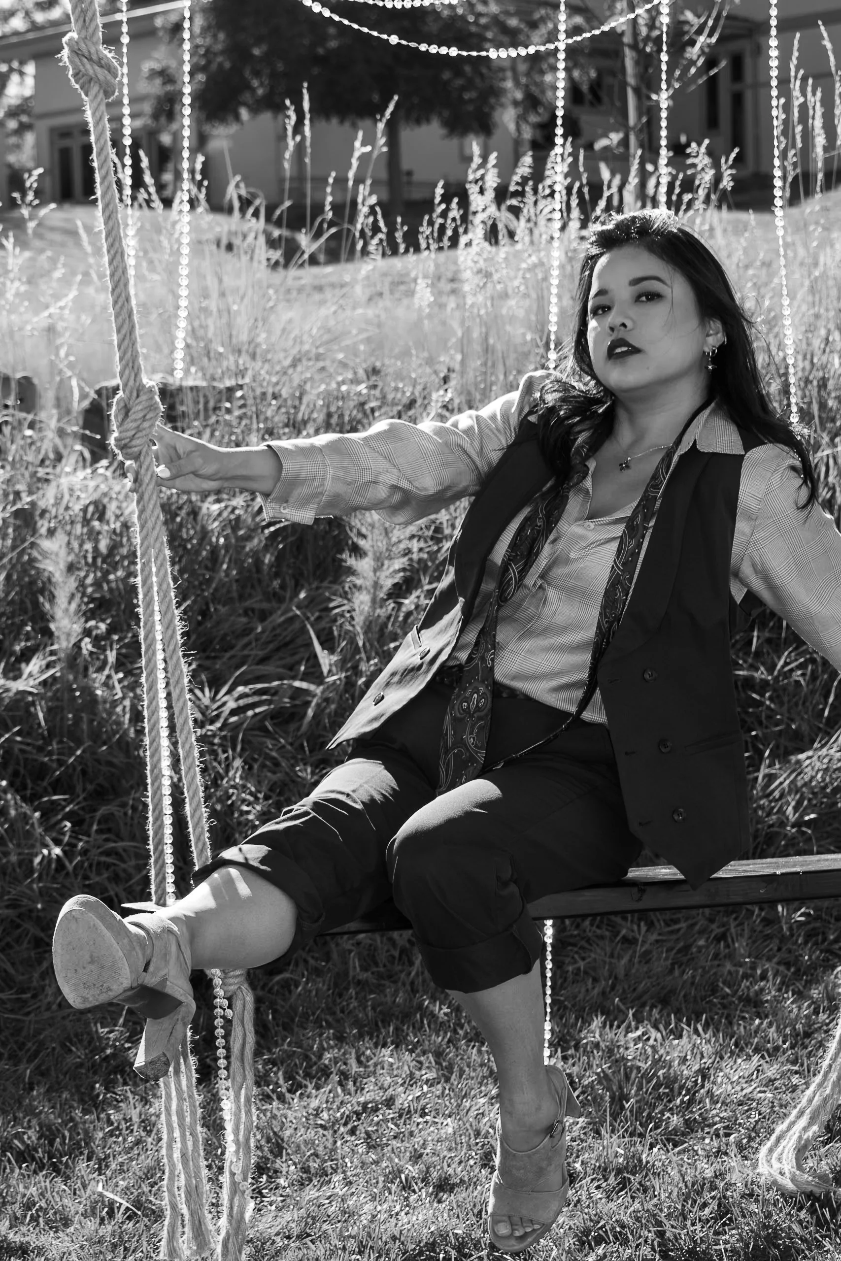 A woman sitting on a swing outdoors, holding the rope with one hand, wearing a blazer, patterned scarf, and high heels, in black and white.