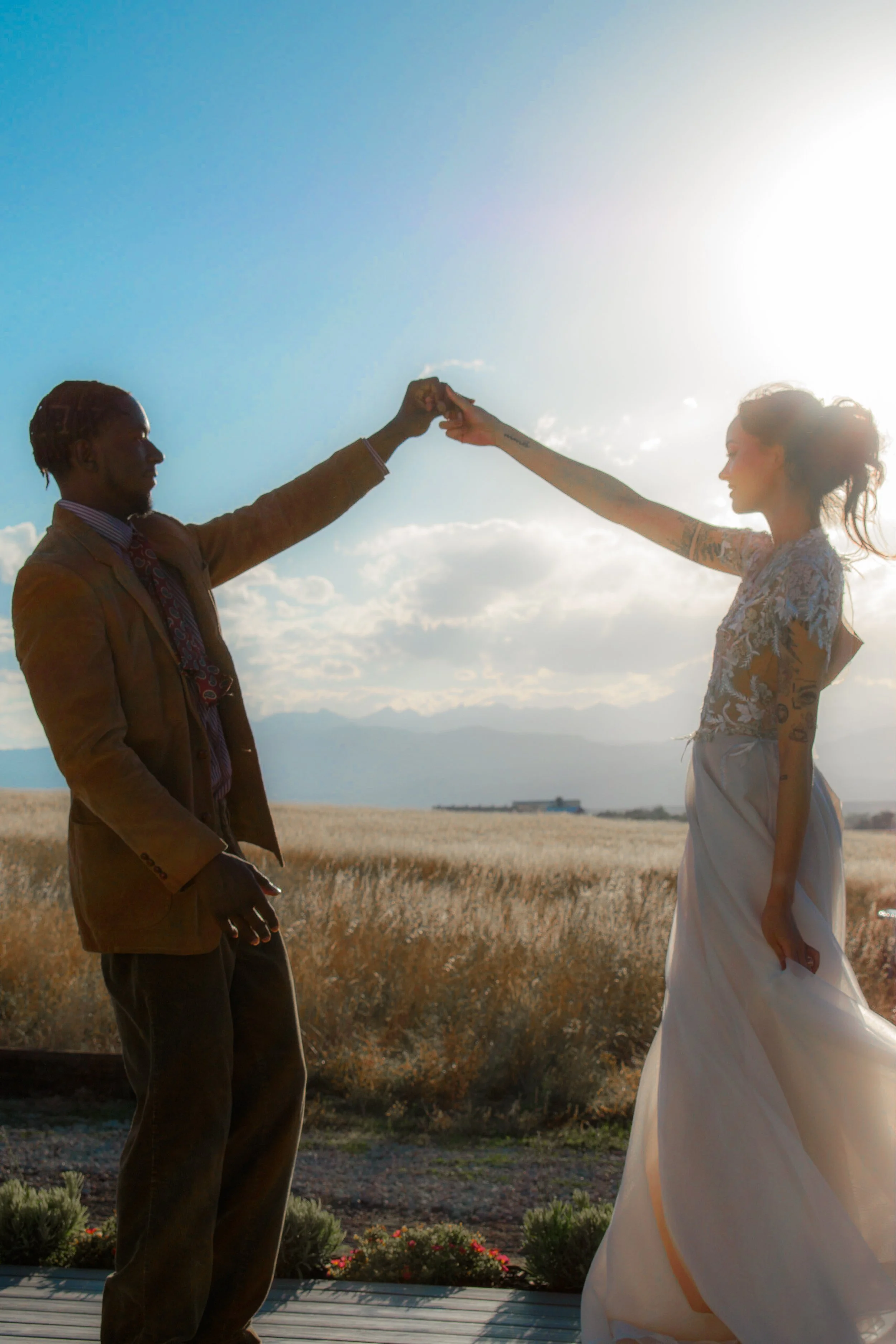 A man and woman holding hands outdoors during sunset, with the woman wearing a white dress and the man in a brown suit, in a desert landscape with mountains in the background.