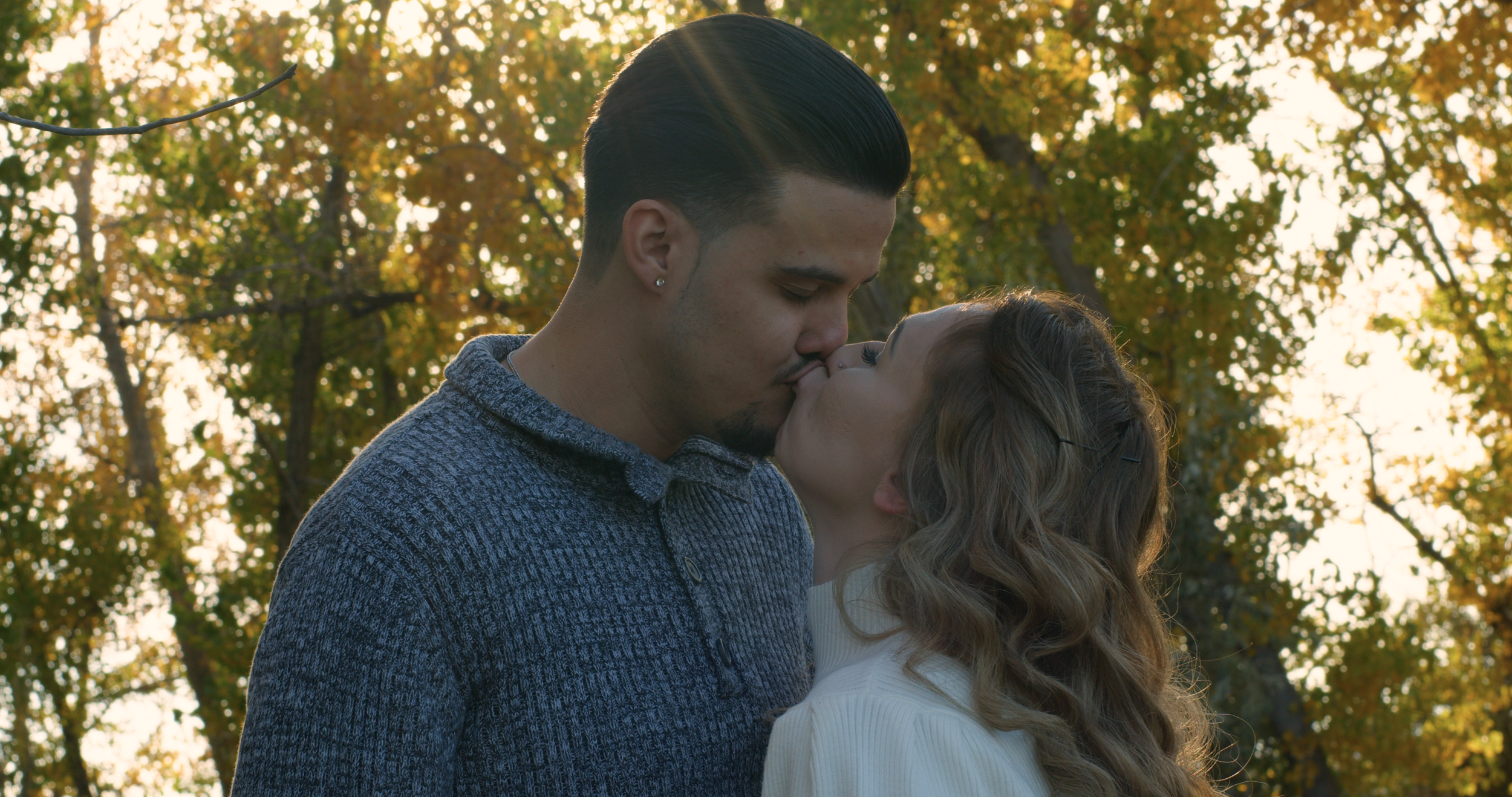 A man and woman kissing outdoors during autumn, with trees showing fall foliage in the background.