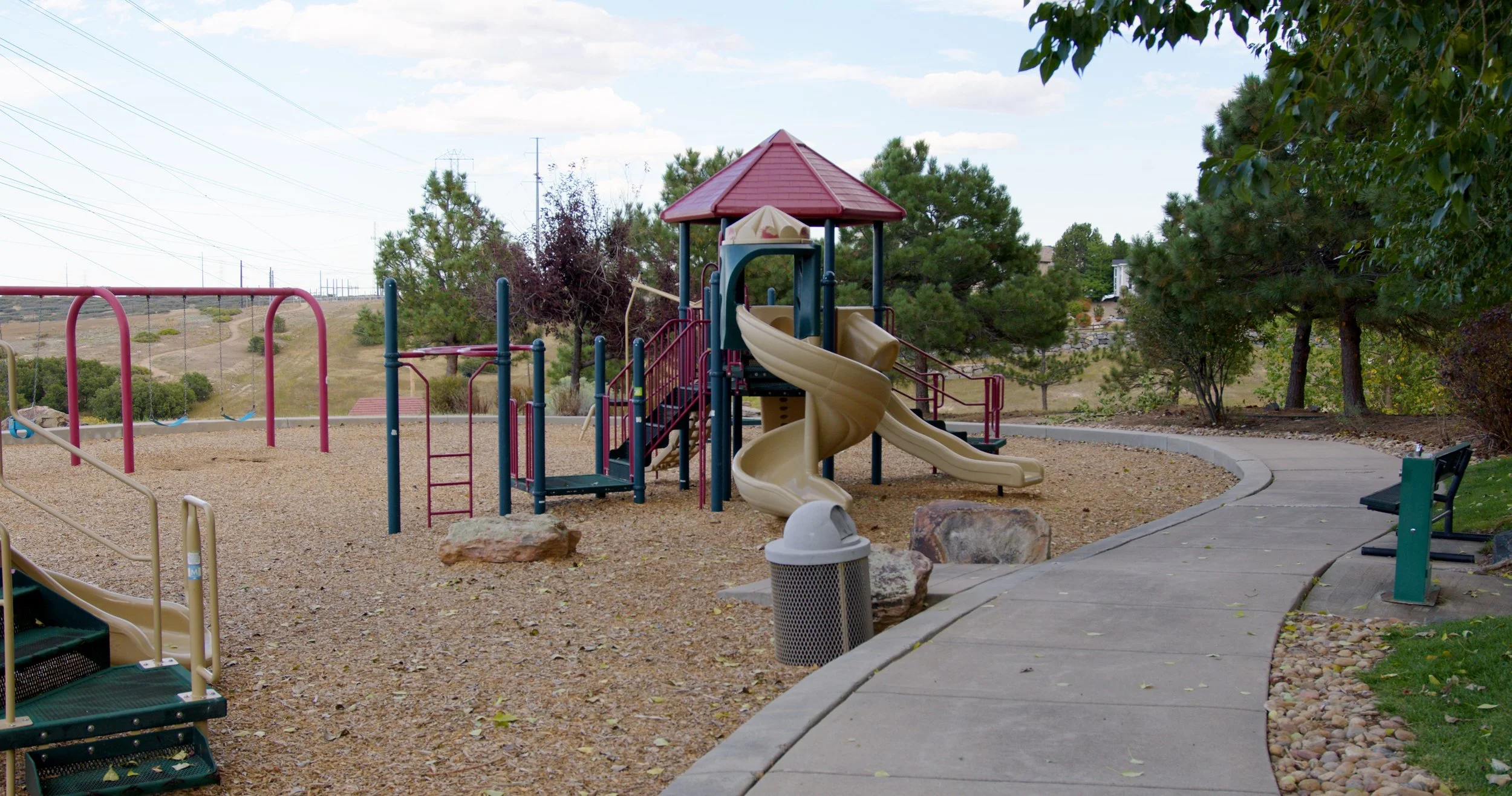Children's playground with slides, swings, and climbing structures surrounded by trees and a curved walking path in a park setting.
