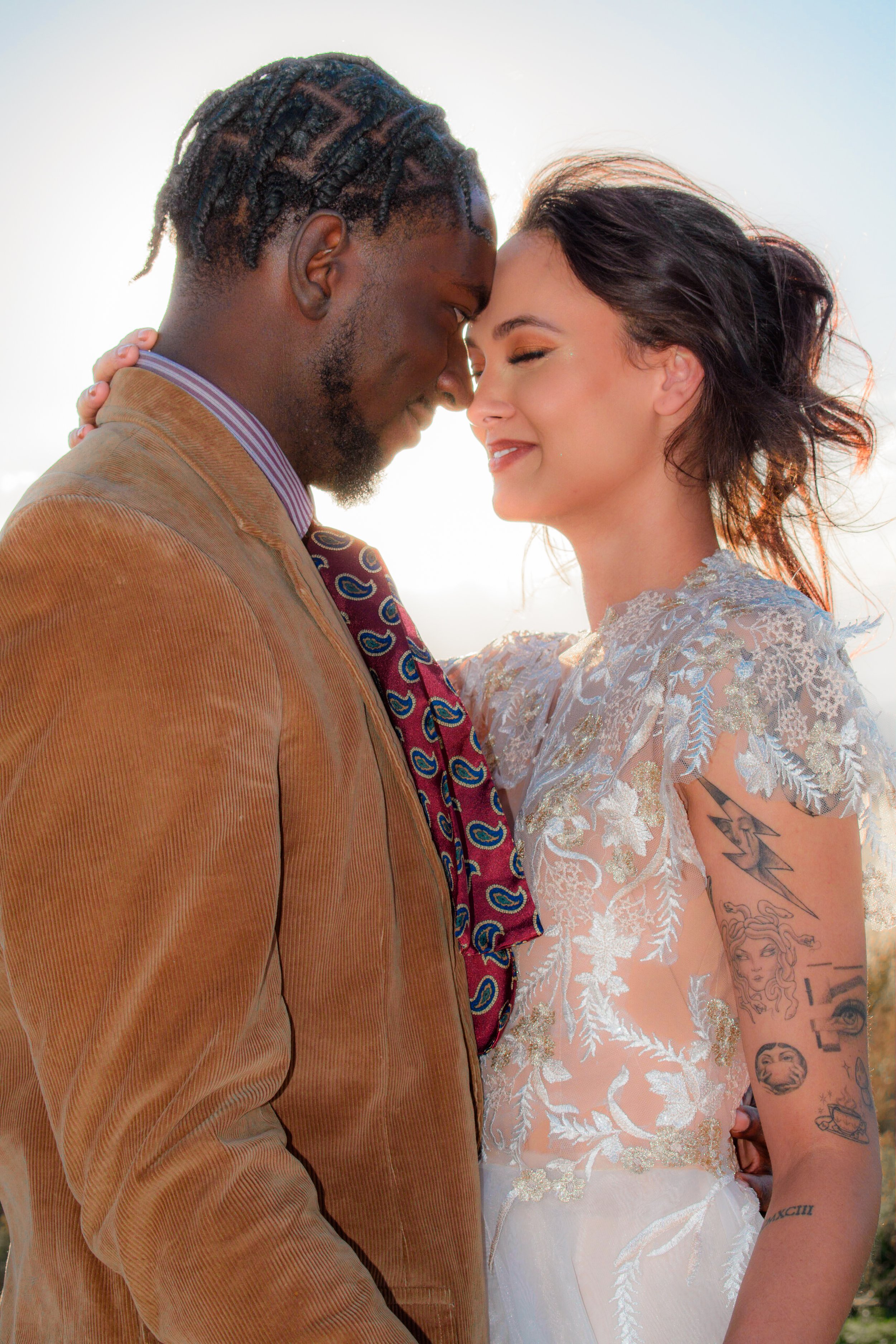 A couple with their foreheads touching, smiling, outdoors with bright background. The man has dark skin, styled hair, and is wearing a tan corduroy jacket and a patterned shirt and tie. The woman has fair skin, dark wavy hair, tattoos on her arm, and